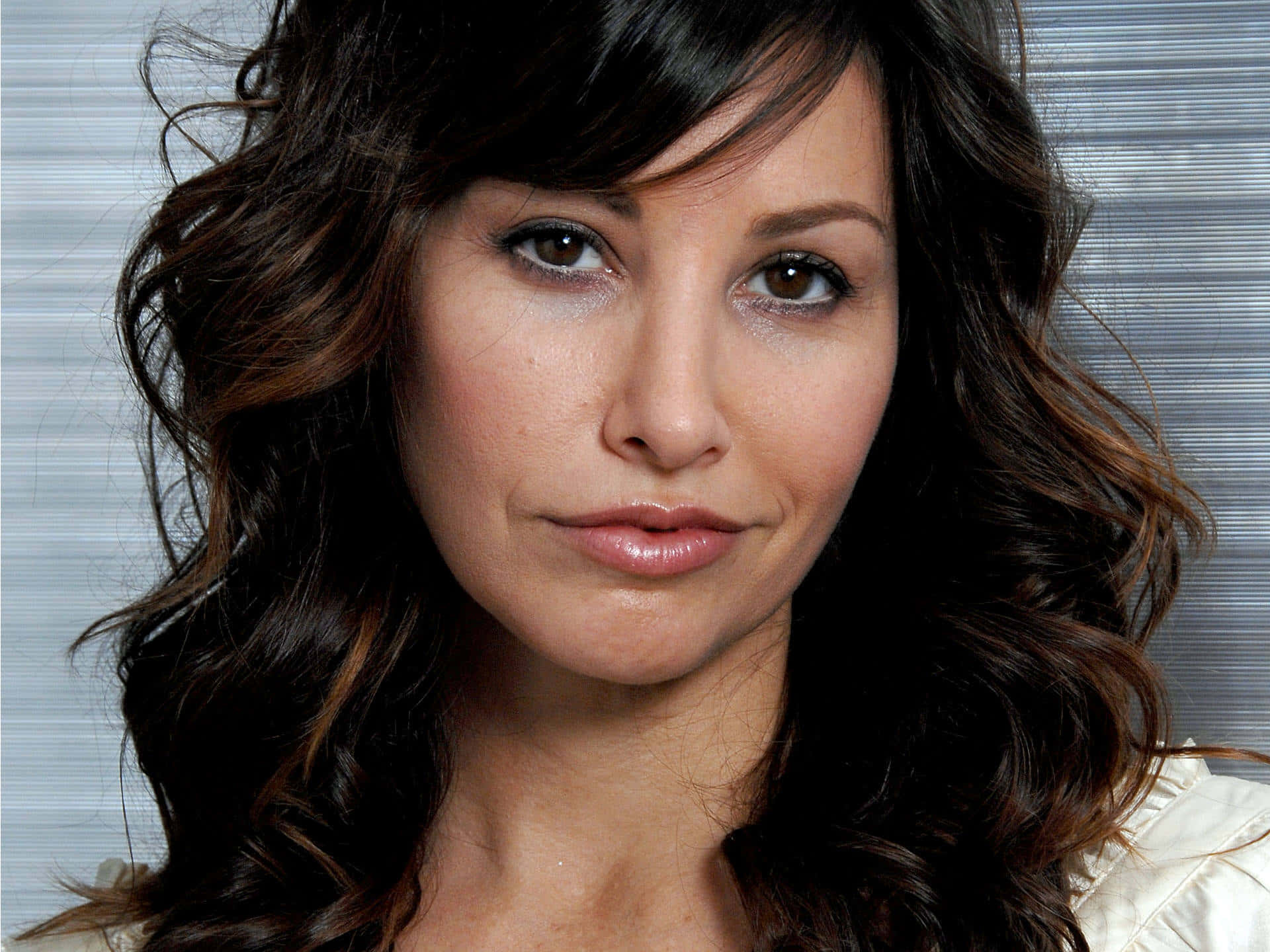 Gina Gershon 桌布 壁紙 — 一位擁有波浪深色頭髮和柔和妝容的女性，在條紋背景前的特寫肖像。