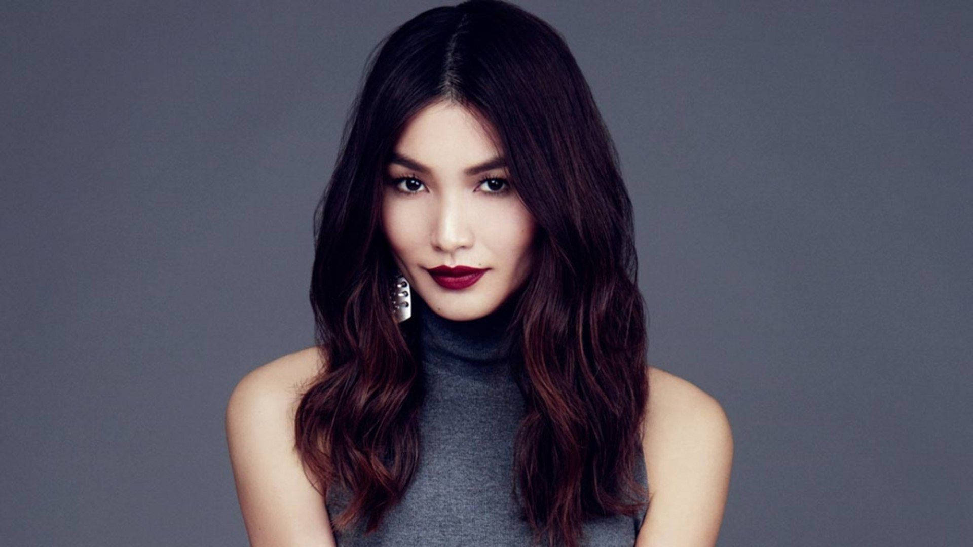 Gemma Chan 桌布 壁紙 — 一位擁有波浪深色頭髮和鮮豔紅唇的女性，在柔和的灰色背景下直視鏡頭。
