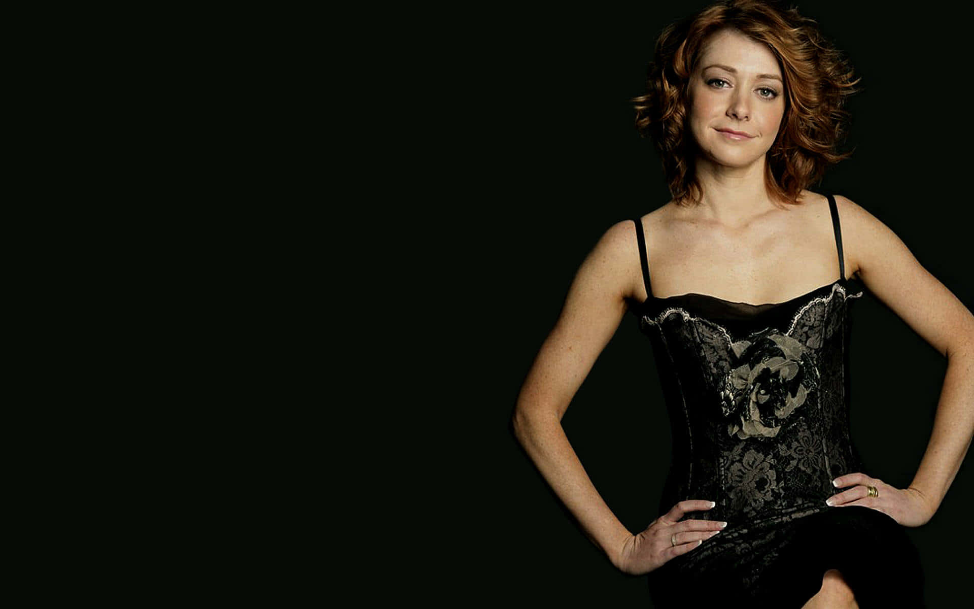 Alyson Hannigan 桌布 壁紙 — 一位擁有波浪赤褐色頭髮的女性，穿著黑色蕾絲緊身胸衣，在純黑色背景前自信地擺姿勢。