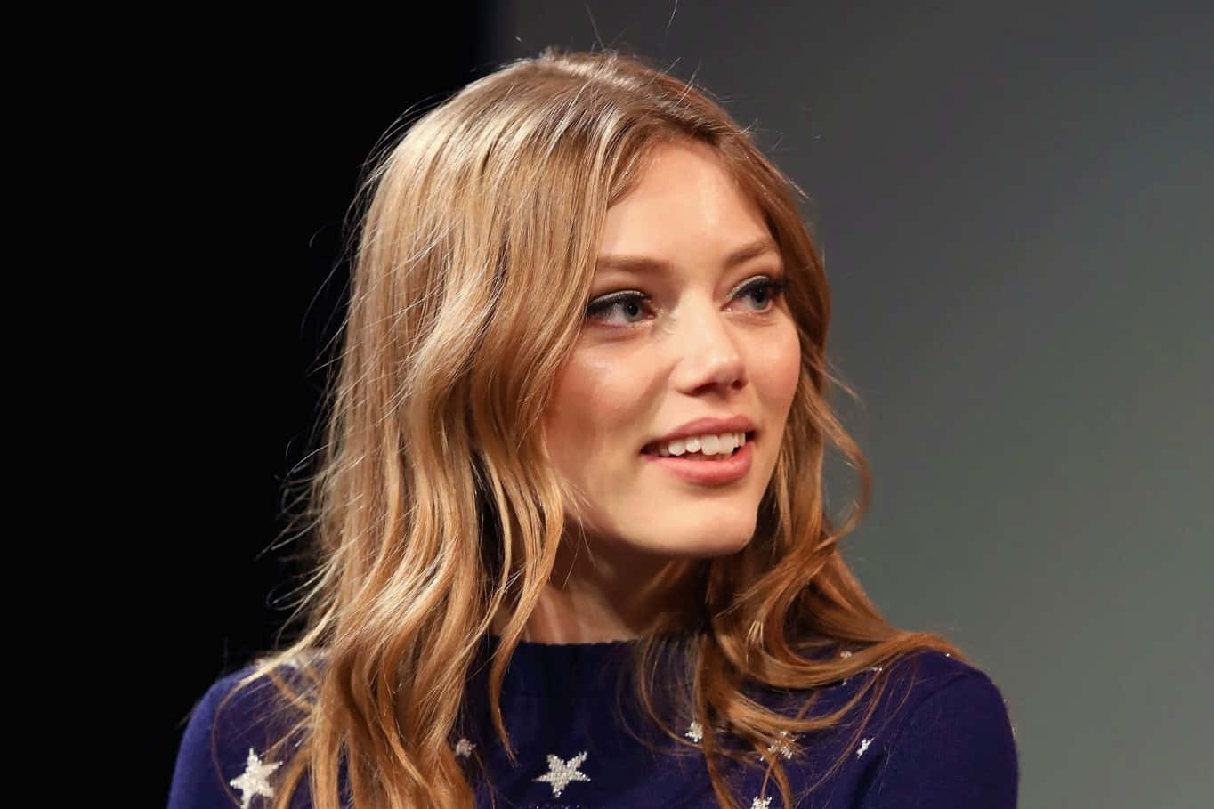Grace Van Dien 桌布 壁紙 — 一位擁有波浪金髮、身穿星星圖案海軍藍毛衣的女性，在深色漸變背景前溫柔微笑。