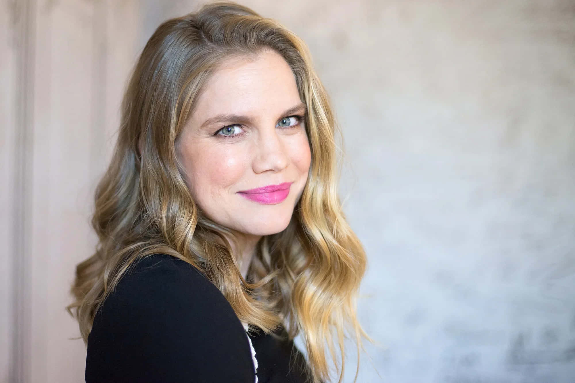 anna chlumsky 桌布 壁紙 — 一位擁有波浪金髮和亮粉紅色唇膏的女性，在帶有紋理的中性色牆壁前溫柔微笑。