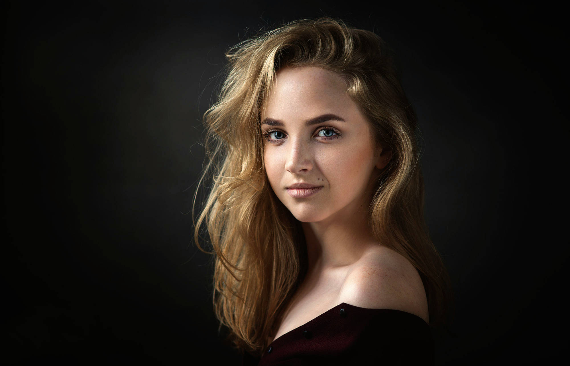 Young Models 桌布 壁紙 — 一位擁有波浪金髮和引人注目的藍眼睛的年輕女性，在深色背景下穿著深紅色露肩上衣，溫柔地凝視著。