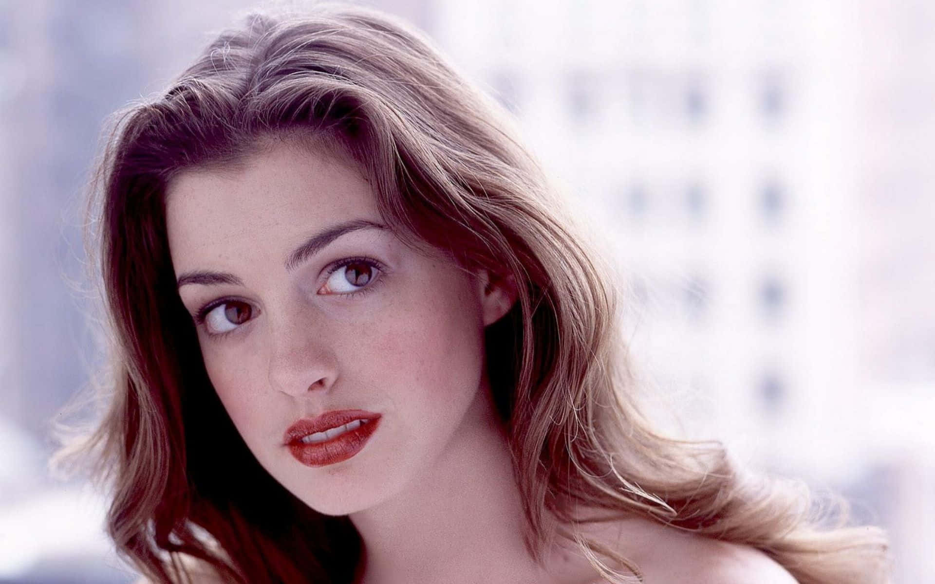 Anne Hathaway 桌布 壁紙 — 一位擁有波浪金髮和紅唇的女性特寫肖像，在模糊的城市景觀背景下沉思地凝視。