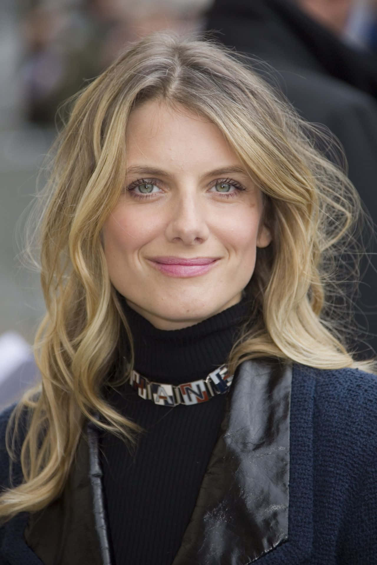 Mélanie Laurent 桌布 壁紙 — 一位擁有波浪金髮和綠色眼睛的女性，穿著黑色高領毛衣和圖案頸鍊，溫柔微笑的特寫肖像。