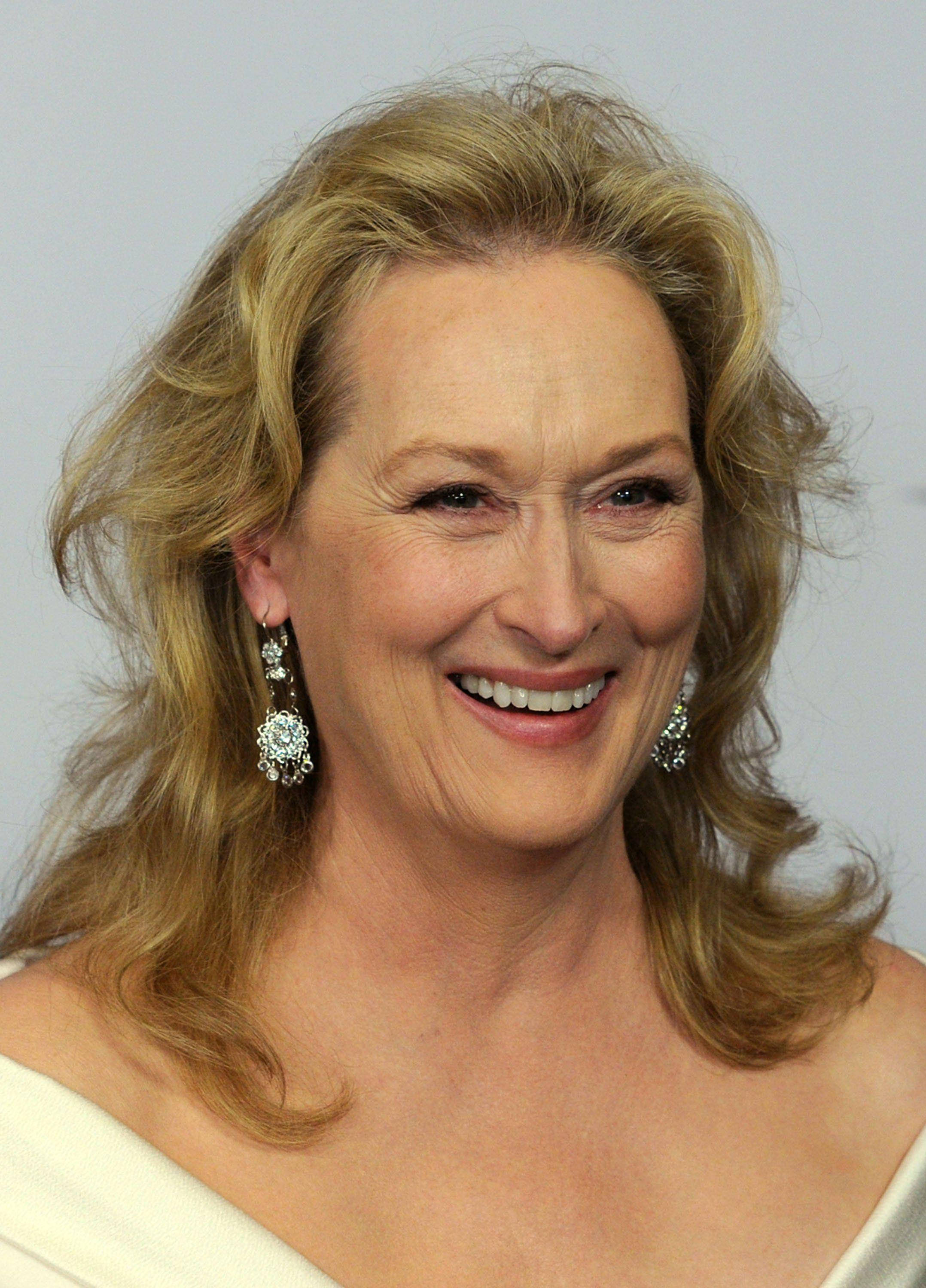 Meryl Streep 桌布 壁紙 — 一位擁有波浪金髮和閃亮耳環的容光煥發的女性，在純色背景前溫暖地微笑。