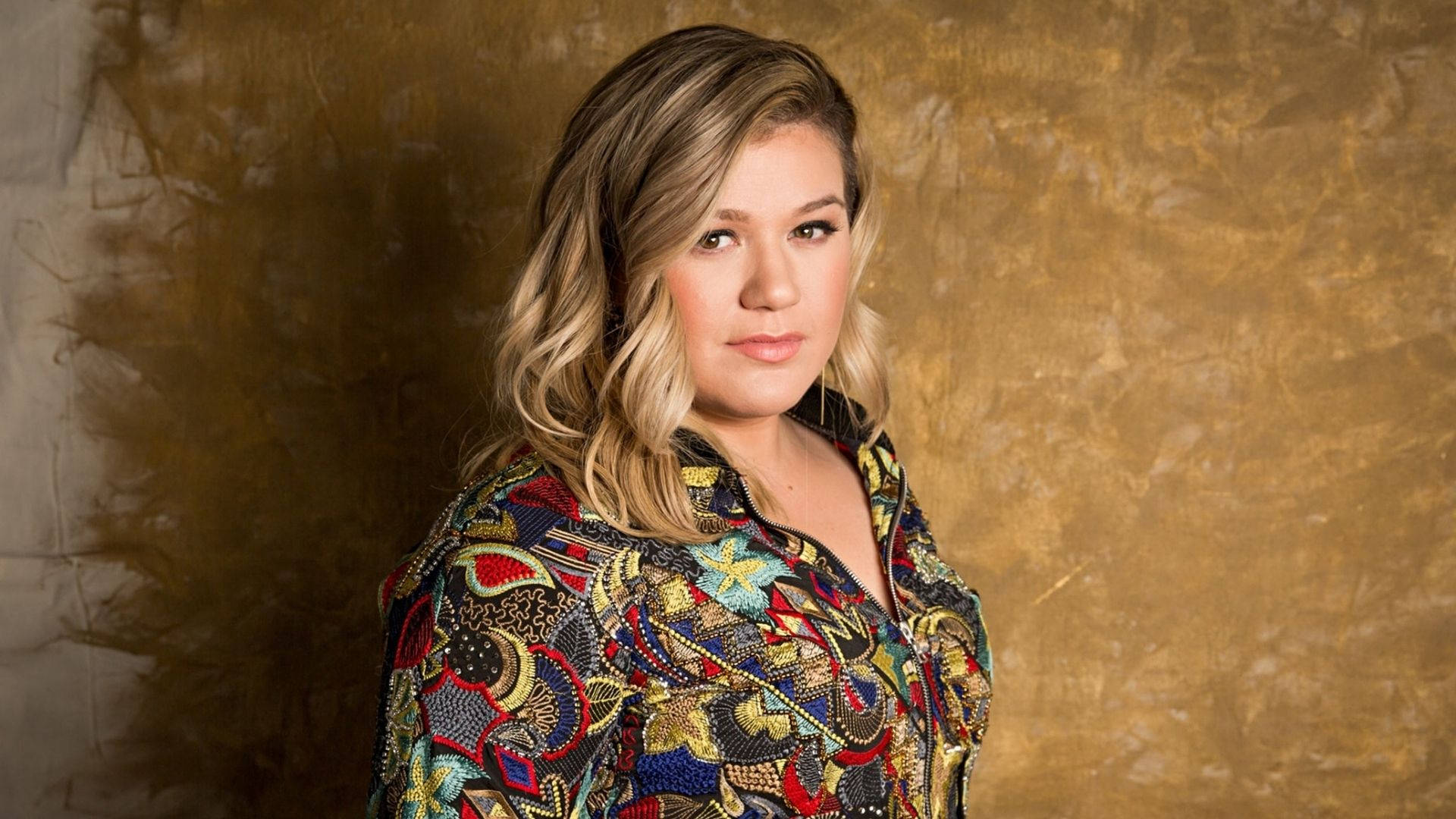 Kelly Clarkson 桌布 壁紙 — 一位擁有波浪金髮的女性，穿著色彩繽紛的圖案襯衫，背景是帶有紋理的金色背景。