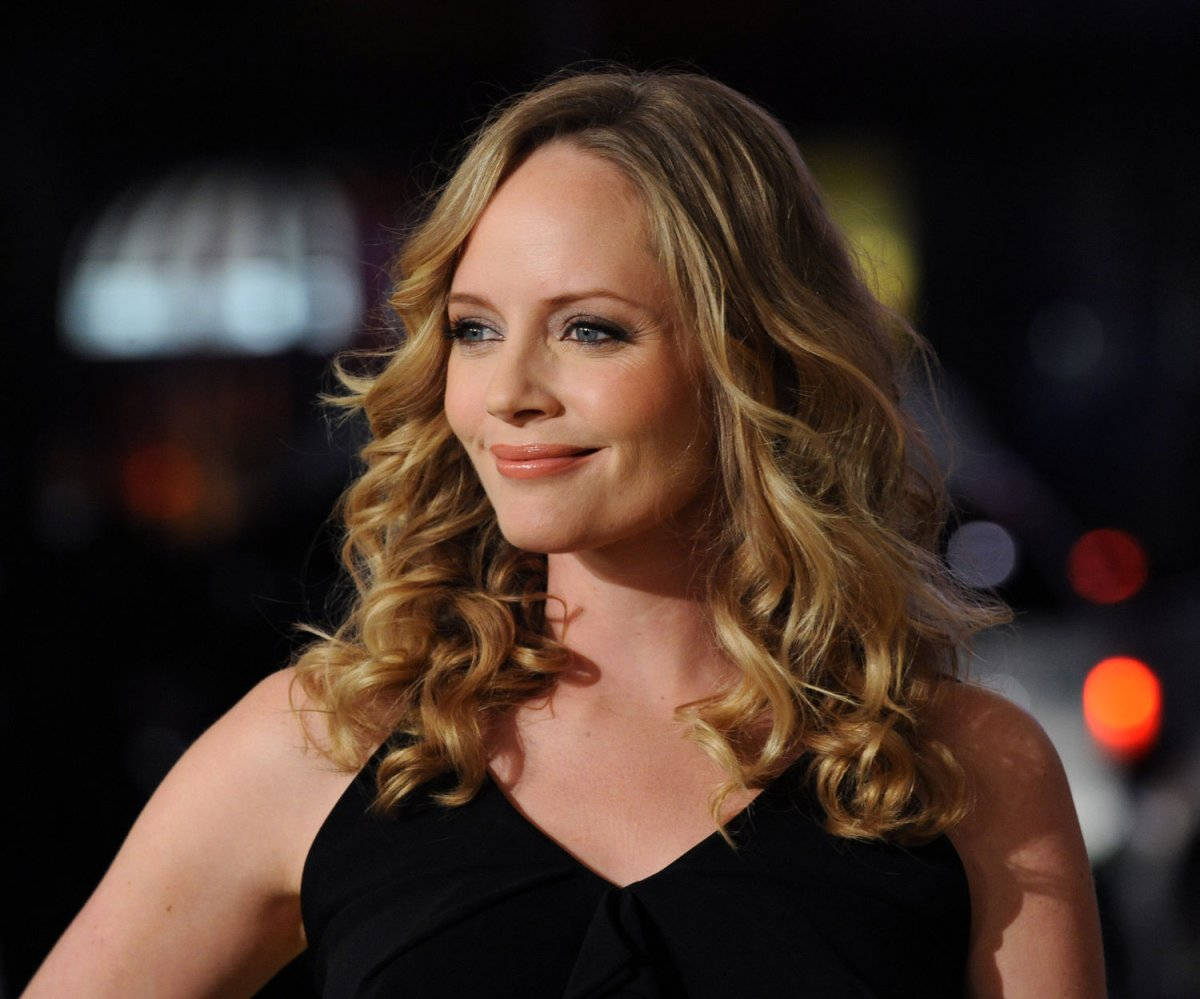 Marley Shelton 桌布 壁紙 — 一位擁有波浪金髮的女性，身穿黑色連衣裙，在模糊的夜間城市背景下溫柔微笑。