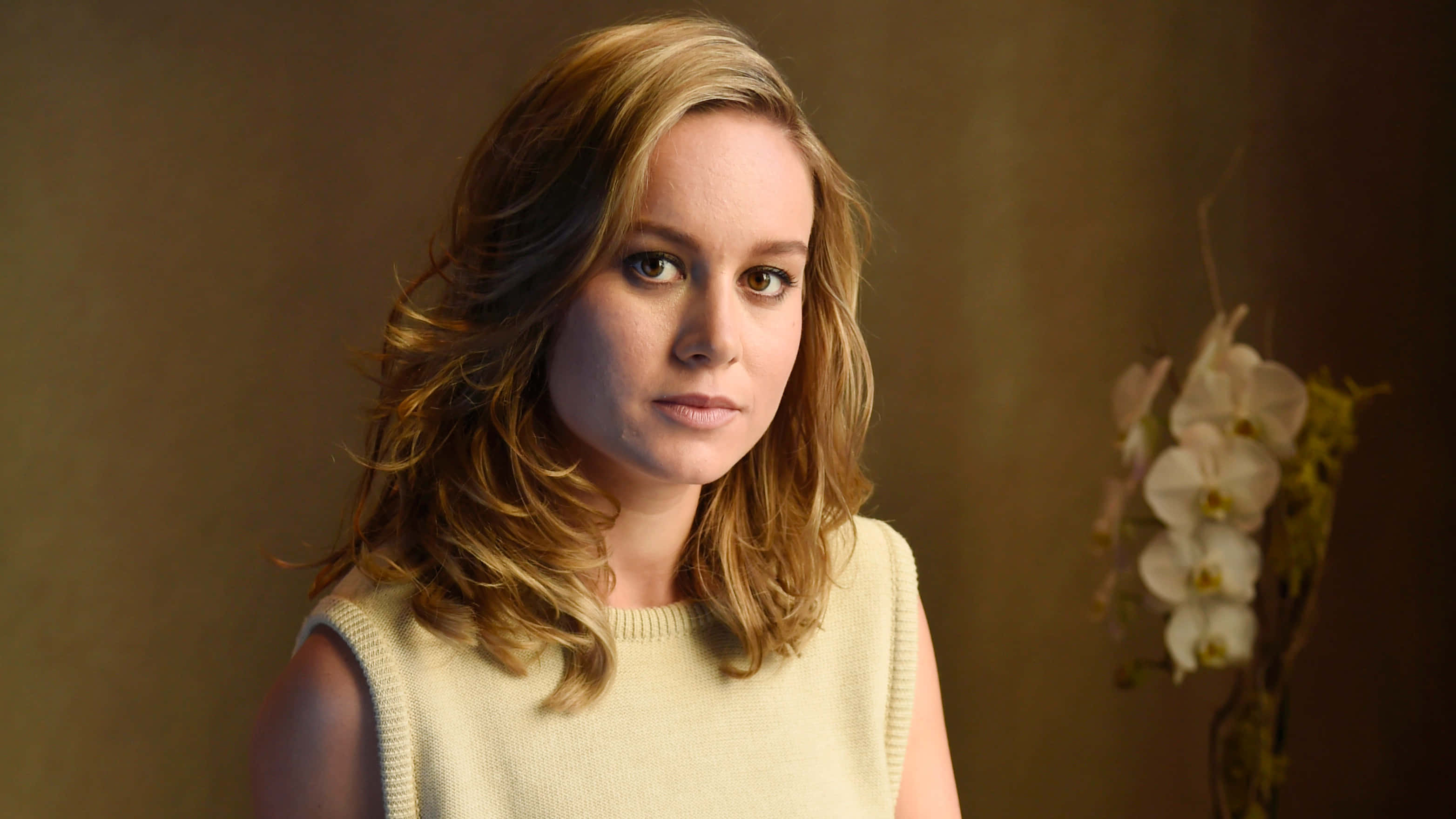 brie larson 桌布 壁紙 — 一位擁有波浪金髮的女性在柔和燈光的攝影棚中，旁邊有白色蘭花，直視鏡頭。