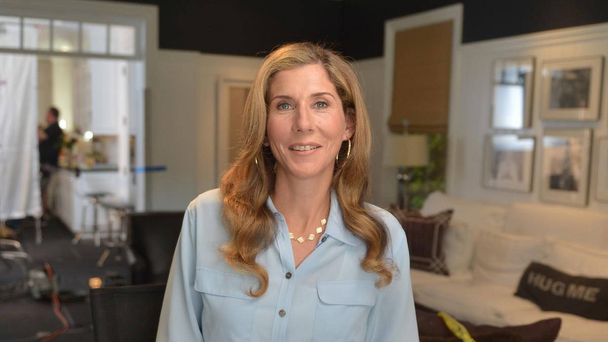 Monica Seles 桌布 壁紙 — 一位擁有波浪金髮的女性在裝潢時尚的客廳中溫暖微笑，客廳內有裱框藝術品和一個「HUG ME」抱枕。