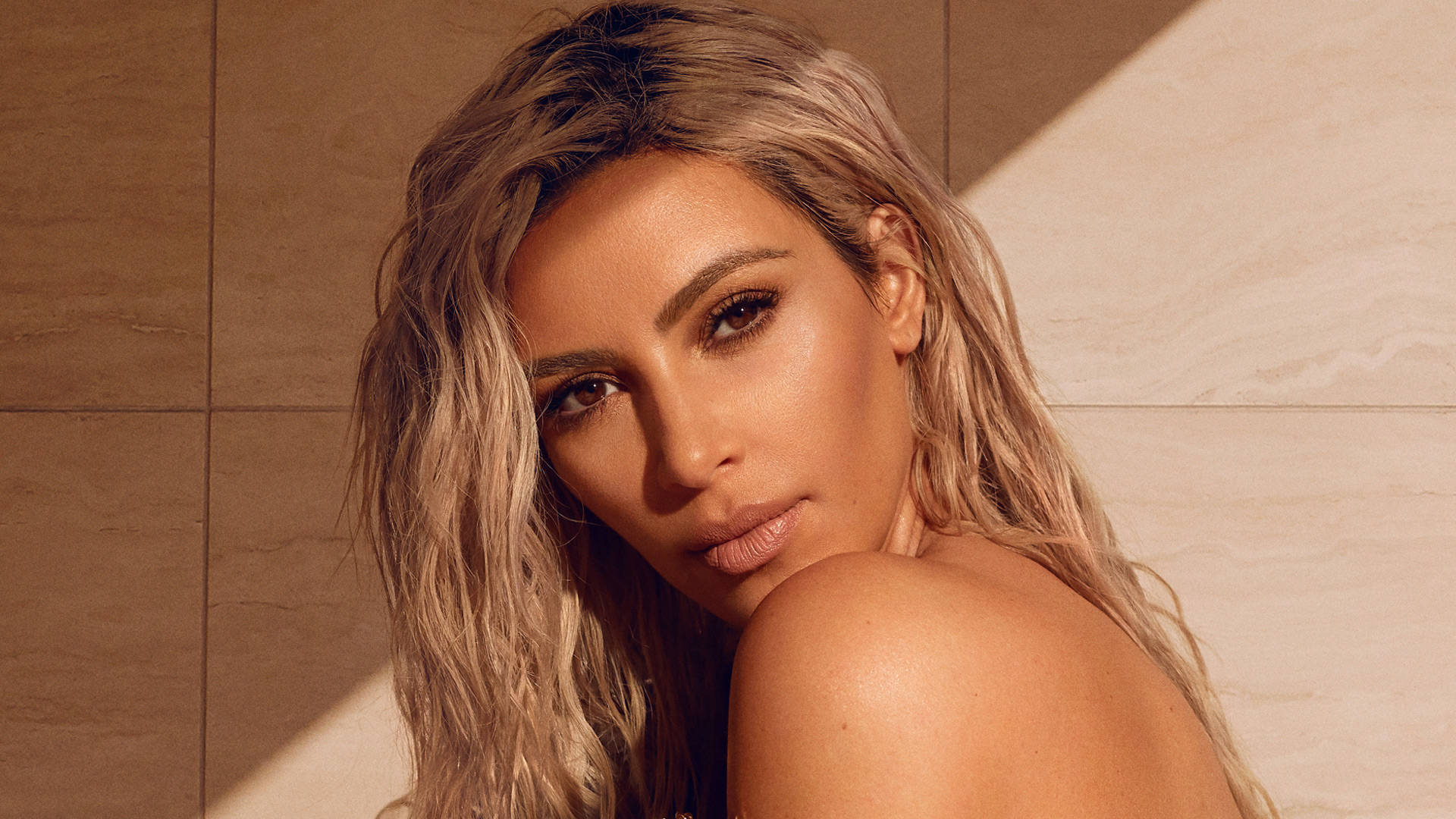 Kim Kardashian 桌布 壁紙 — 一位擁有波浪金髮的女性直視鏡頭，背景是暖色調的瓷磚牆，柔和陽光投射出幾何陰影。