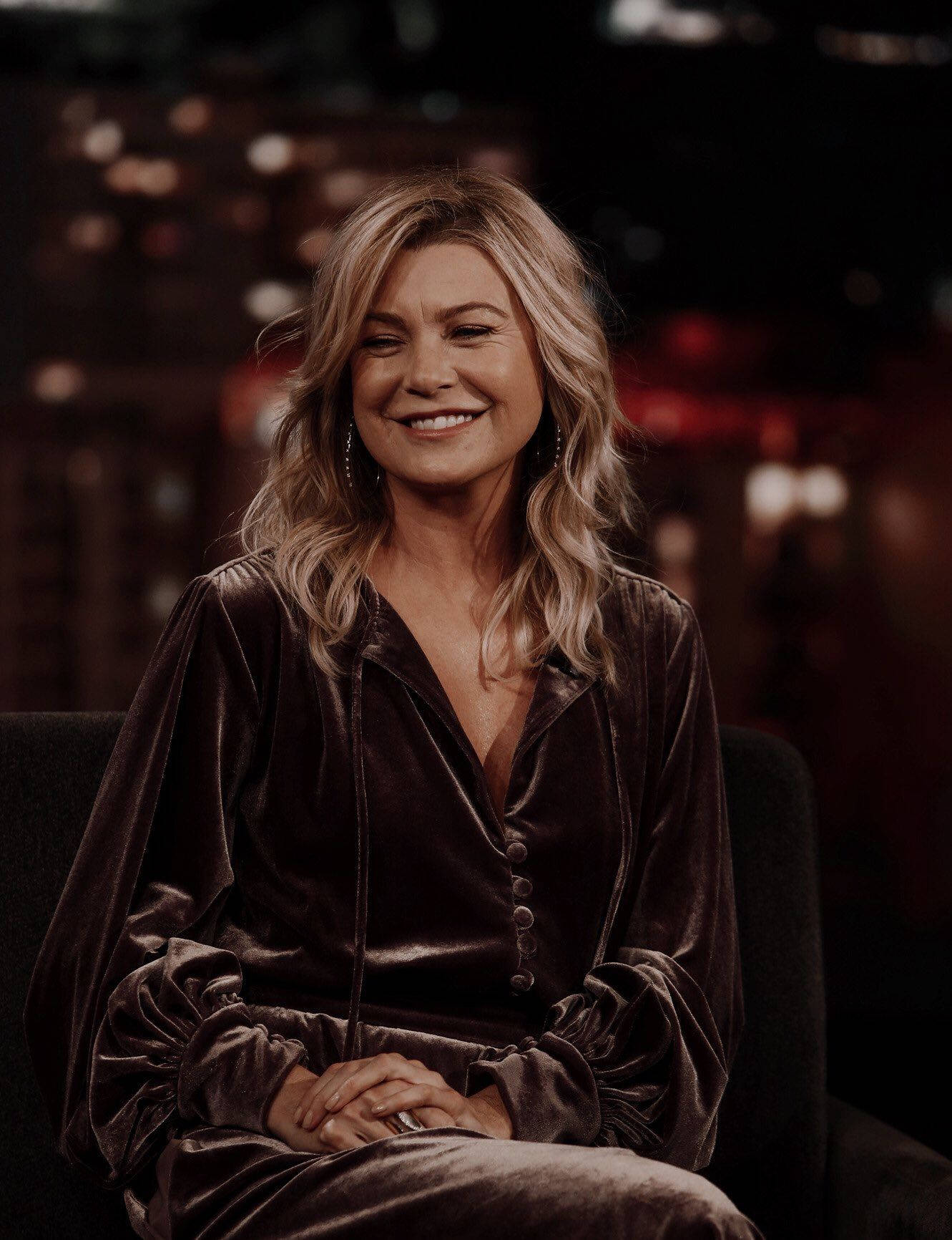 Ellen Pompeo 桌布 壁紙 — 一位擁有波浪金髮的女性穿著深色天鵝絨服裝，在模糊的城市夜景背景下溫暖地微笑。