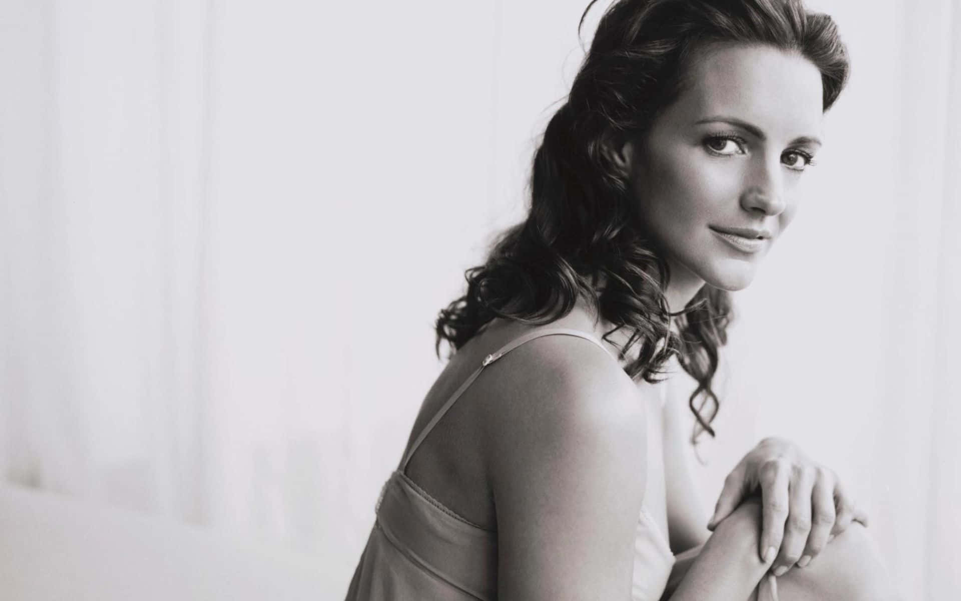 kristin davis 桌布 壁紙 — 一位擁有波浪長髮的女性，坐在明亮的白色窗簾背景前，在黑白肖像中溫柔微笑。