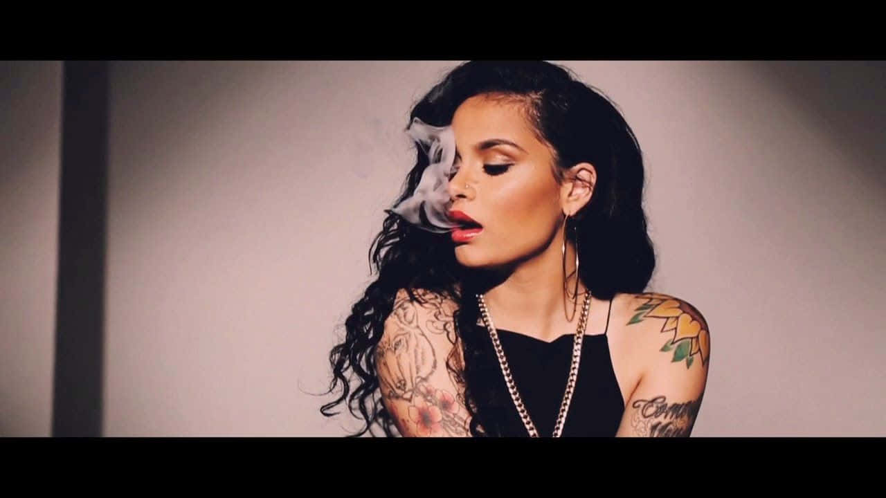 kehlani 桌布 壁紙 — 一位擁有深色捲髮和刺青的女性，穿著黑色上衣和鏈條項鍊，靠著素色牆壁呼出煙霧。