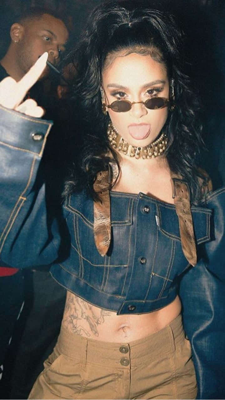 kehlani 桌布 壁紙 — 一位擁有深色捲髮和太陽眼鏡的女性，在昏暗的環境中穿著牛仔短上衣和金色頸鏈，伸出舌頭。
