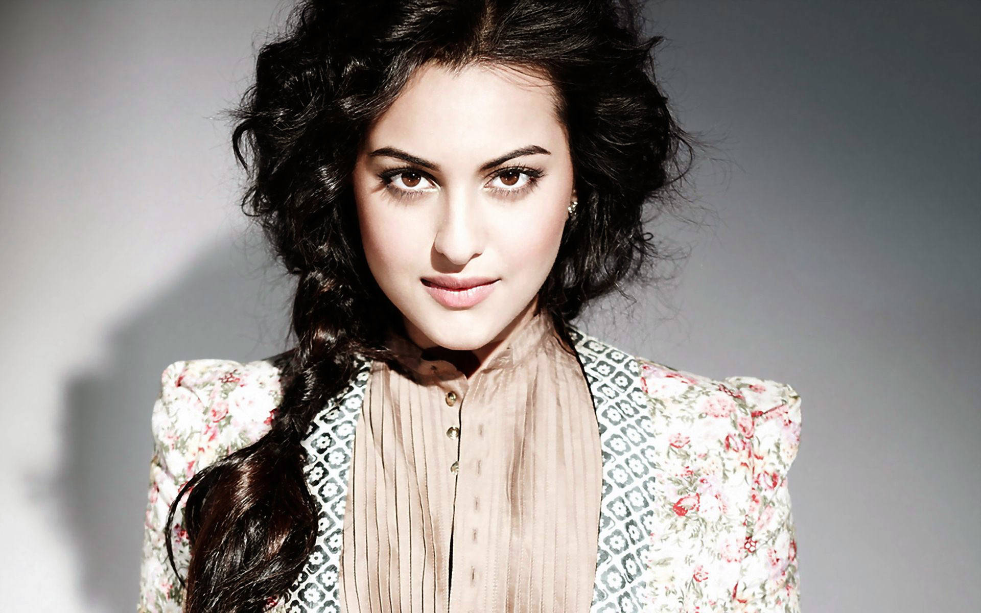 Sonakshi Sinha 桌布 壁紙 — 一位擁有深色捲髮和花卉外套的女性，在柔和的灰色工作室背景下自信地凝視。