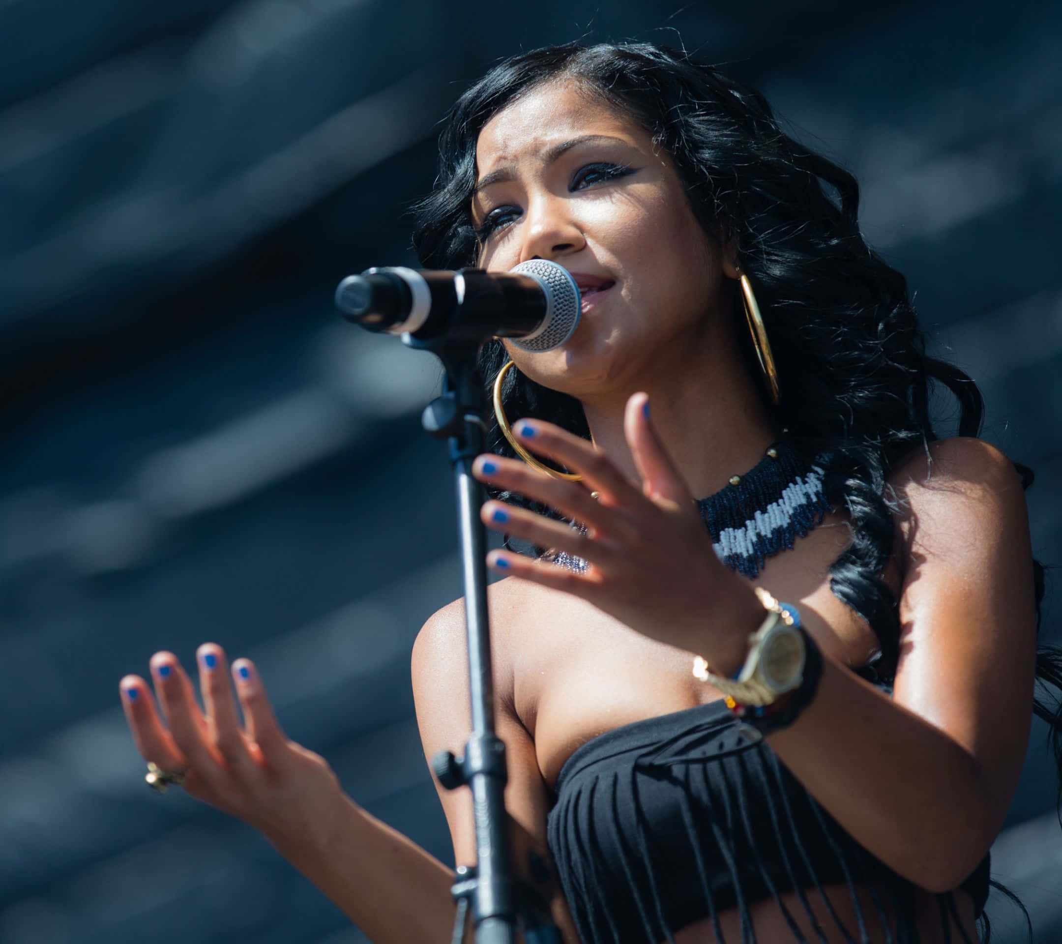 jhene aiko 桌布 壁紙 — 一位擁有深色捲髮和金耳環的女歌手在明亮的日光下熱情地對著麥克風演唱。