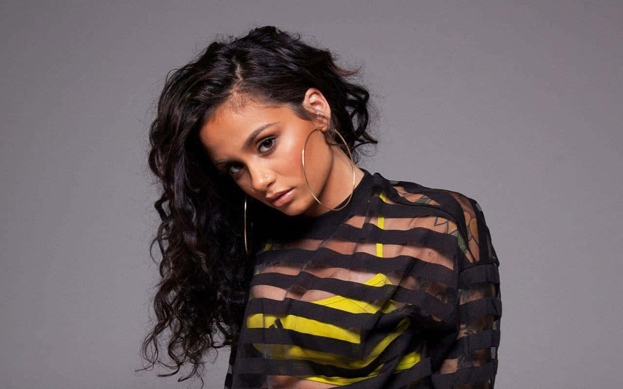 kehlani 桌布 壁紙 — 一位擁有深色捲髮和金色耳環的女性，在灰色背景前穿著黑色和黃色條紋的透視上衣。