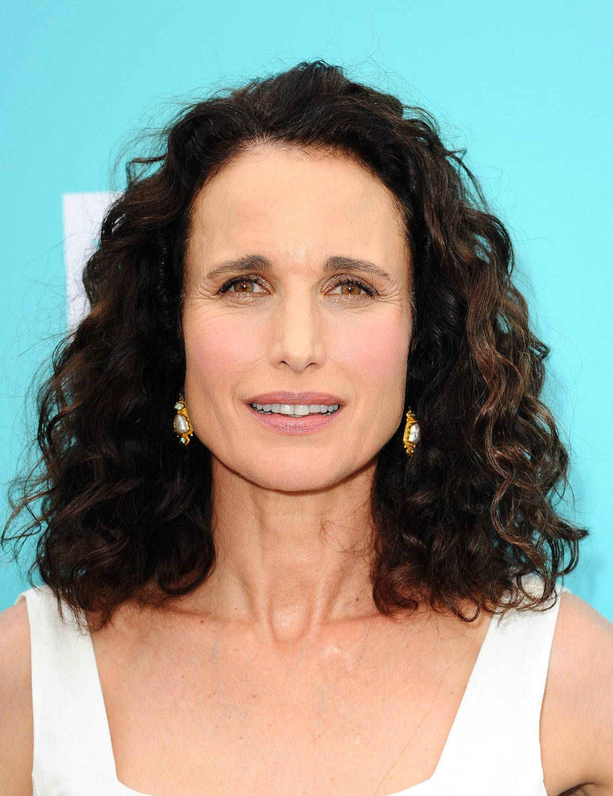 andie macdowell 桌布 壁紙 — 一位擁有深色捲髮和金色耳環的女性在明亮的藍色背景前溫柔地微笑。