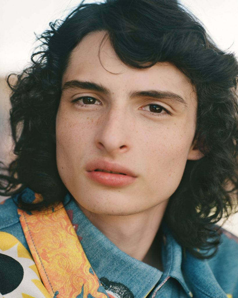 finn wolfhard 桌布 壁紙 — 一位擁有深色捲髮和雀斑的年輕男子，身穿牛仔外套，內搭色彩繽紛的圖案襯衫的 close-up 肖像。