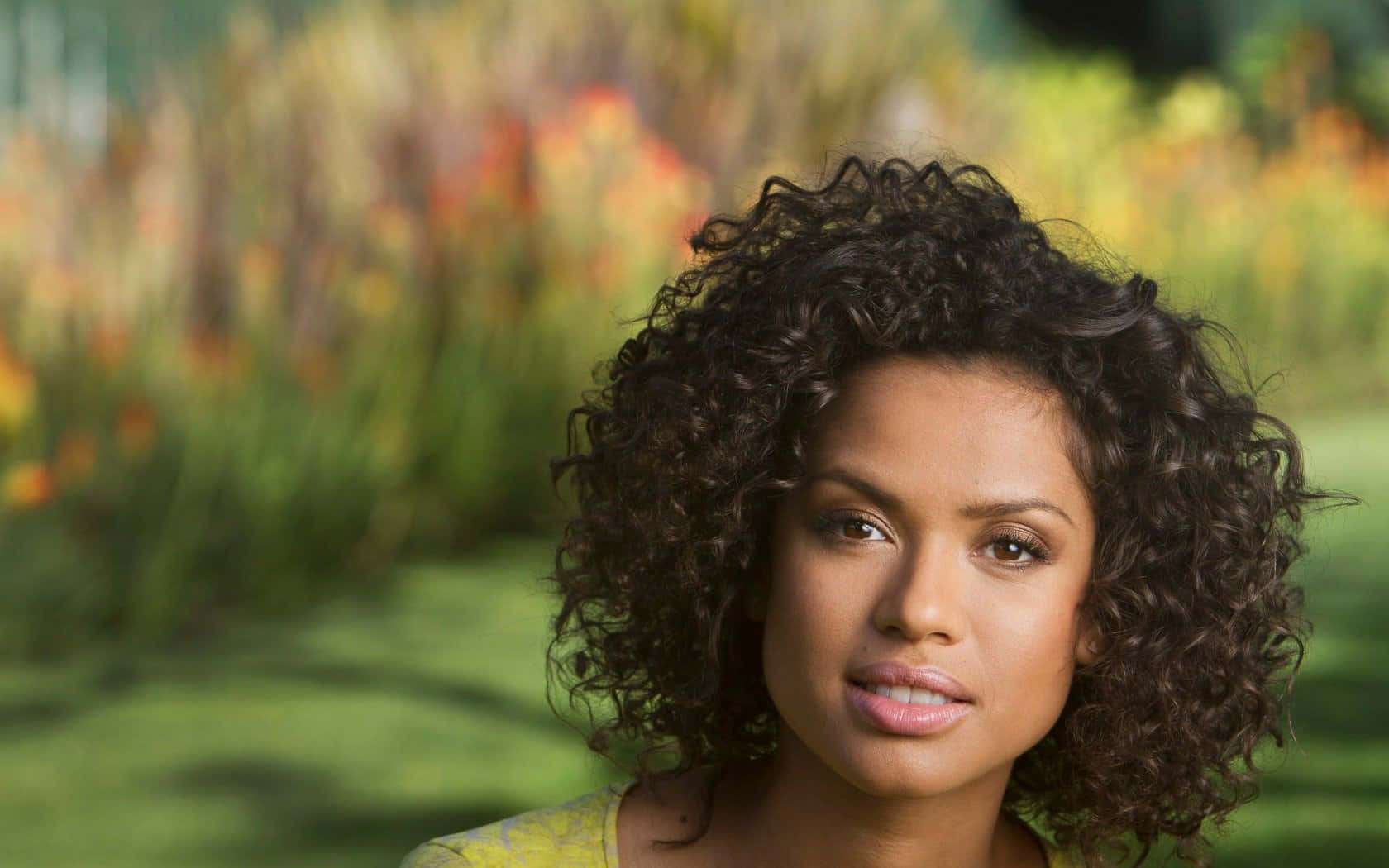 Gugu Mbatha-raw 桌布 壁紙 — 一位擁有深色捲髮的女性，在充滿活力的模糊花園背景下平靜地凝視著鏡頭。