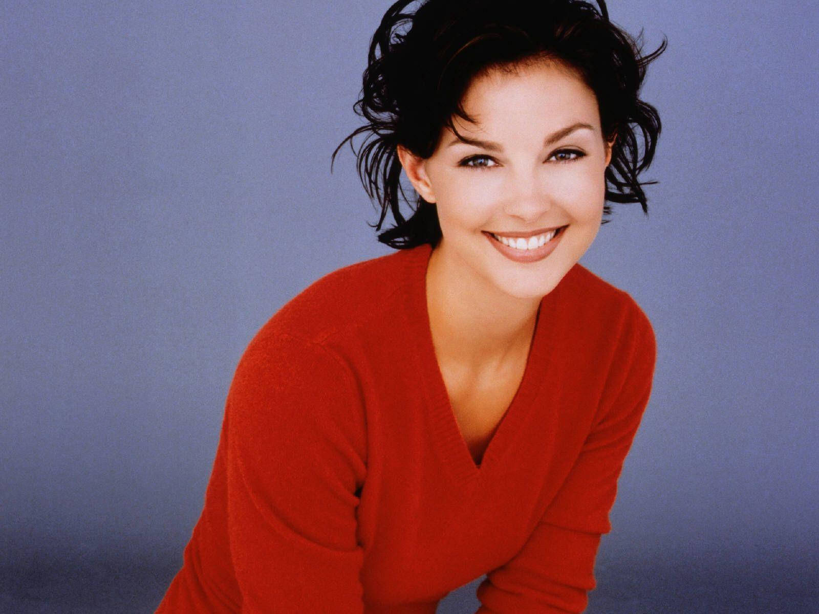 Ashley Judd 桌布 壁紙 — 一位擁有深色捲髮的女性，在柔和的藍色工作室背景前，穿著鮮紅色的毛衣，溫暖地微笑。