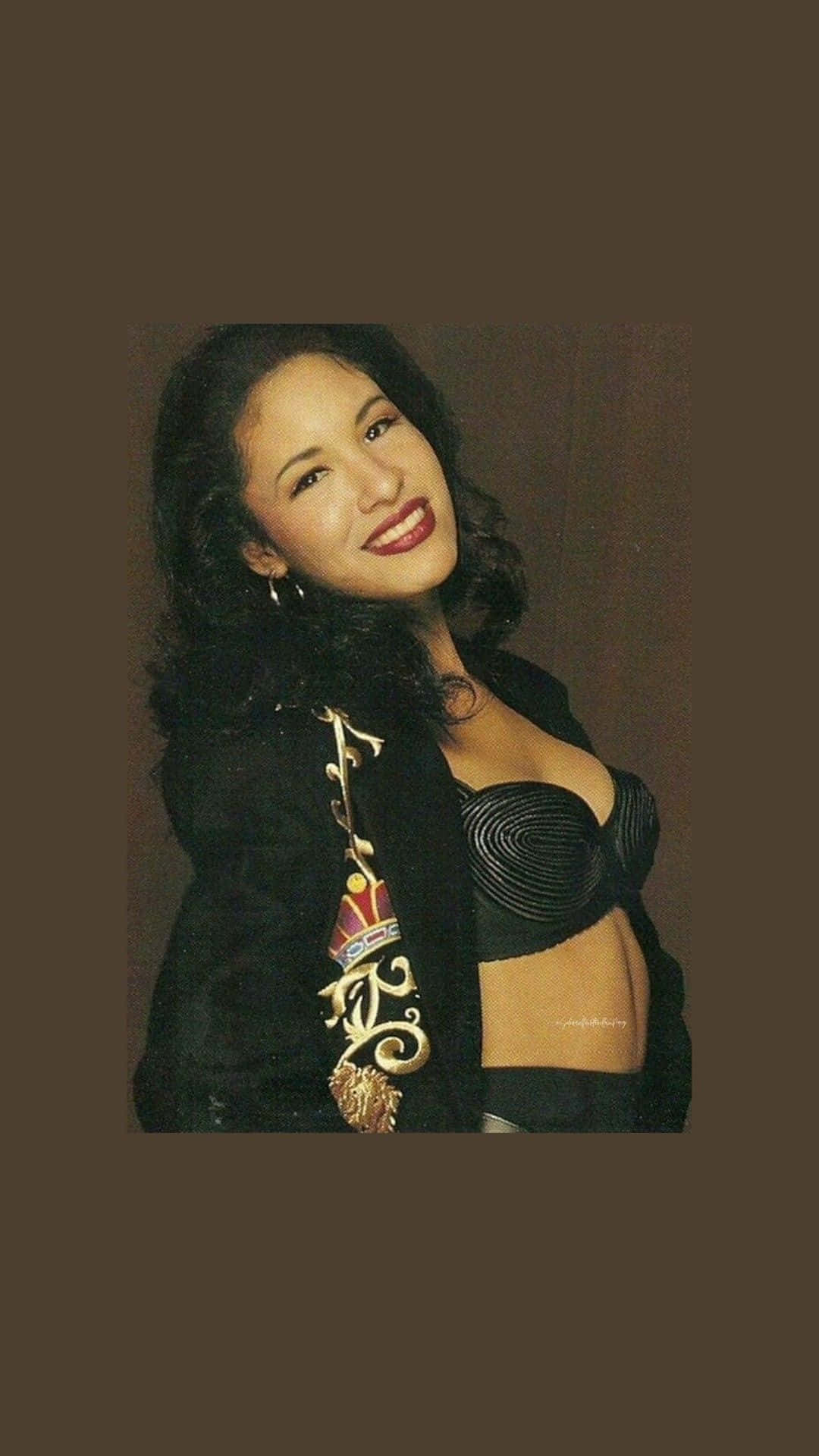 Selena Quintanilla iPhone 桌布 壁紙 — 一位擁有深色捲髮的女性，穿著黑色束腰上衣和帶有華麗金色刺繡的黑色外套，溫暖地微笑。