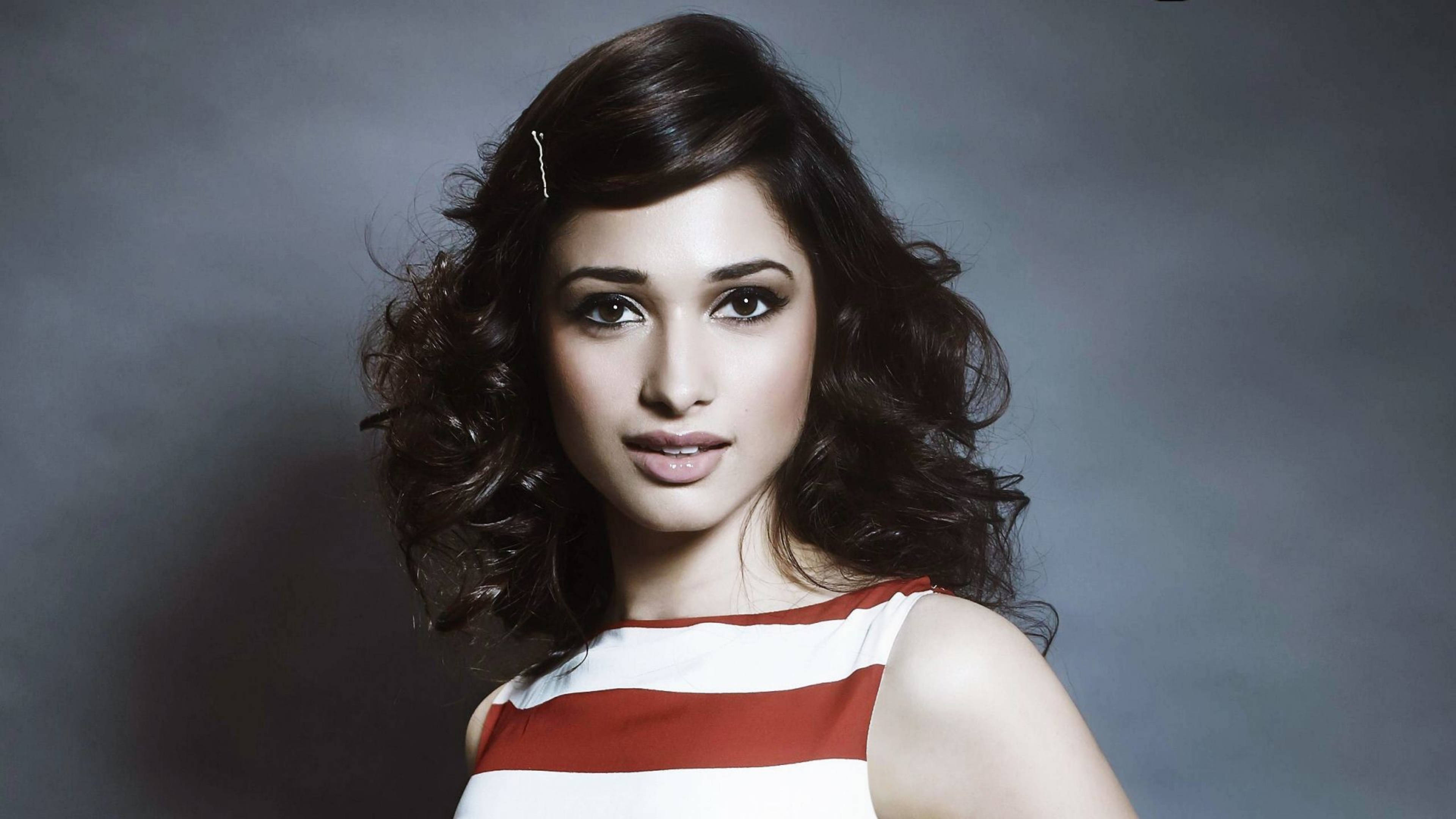 tamannaah bhatia 桌布 壁紙 — 一位擁有深色捲髮的女性，身穿紅白條紋上衣，在柔和的灰色工作室背景前擺姿勢。