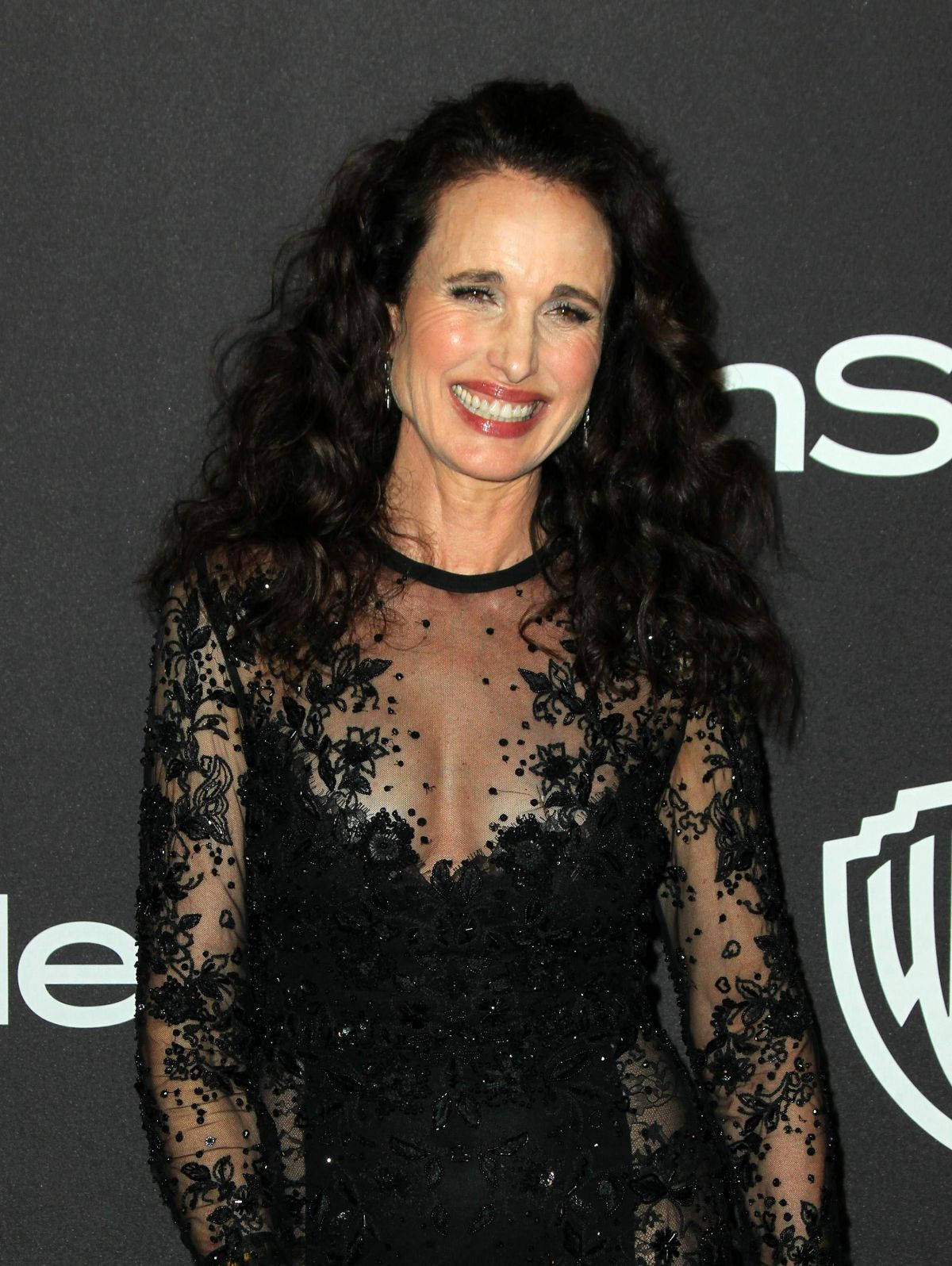 andie macdowell 桌布 壁紙 — 一位擁有深色捲髮的女性在帶有標誌的深色背景前穿著黑色蕾絲連衣裙燦爛微笑。