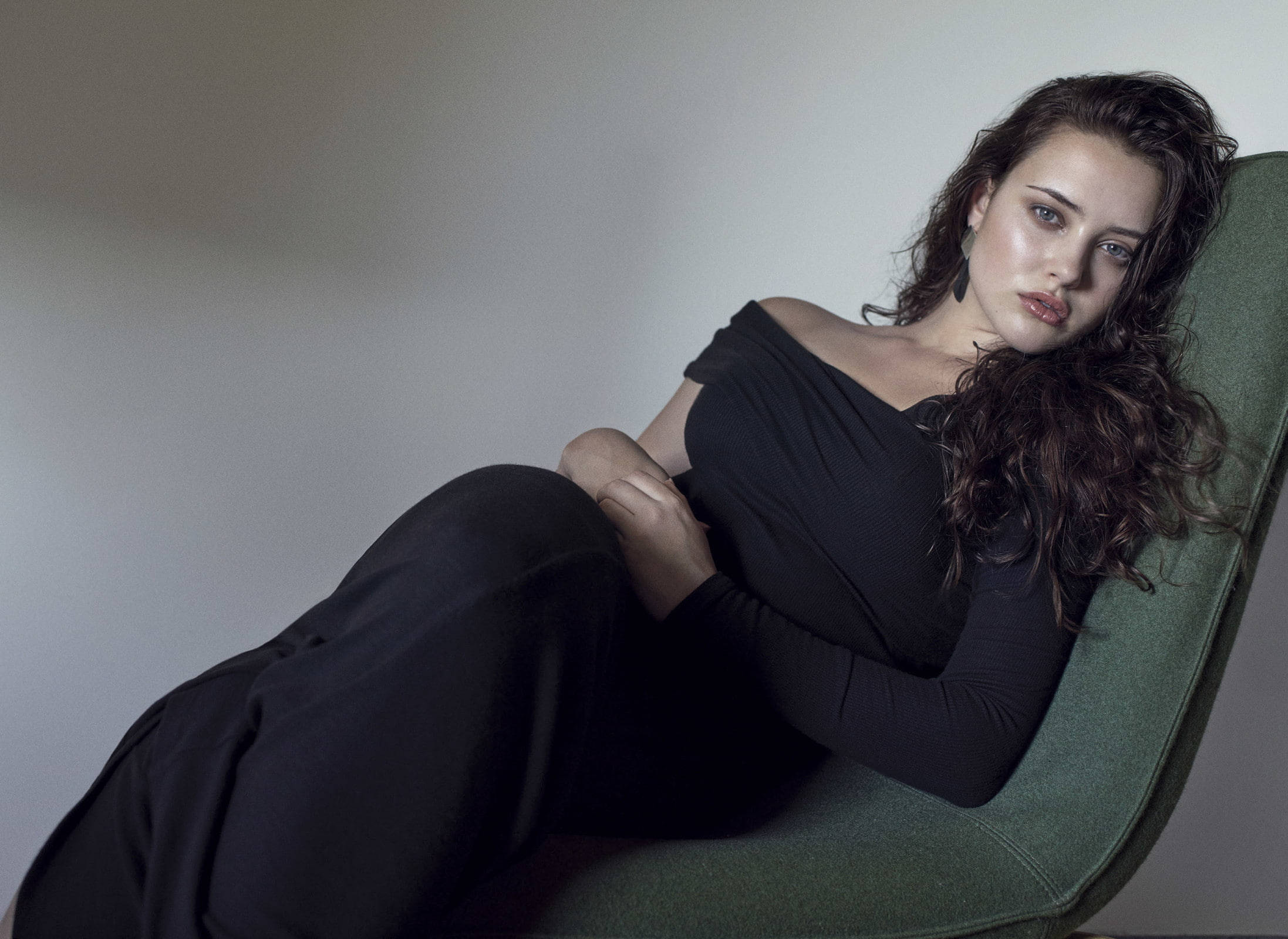 Katherine Langford 桌布 壁紙 — 一位擁有深色捲髮的女性在柔和的攝影棚燈光下，穿著黑色露肩連衣裙，懶洋洋地躺在綠色椅子上。