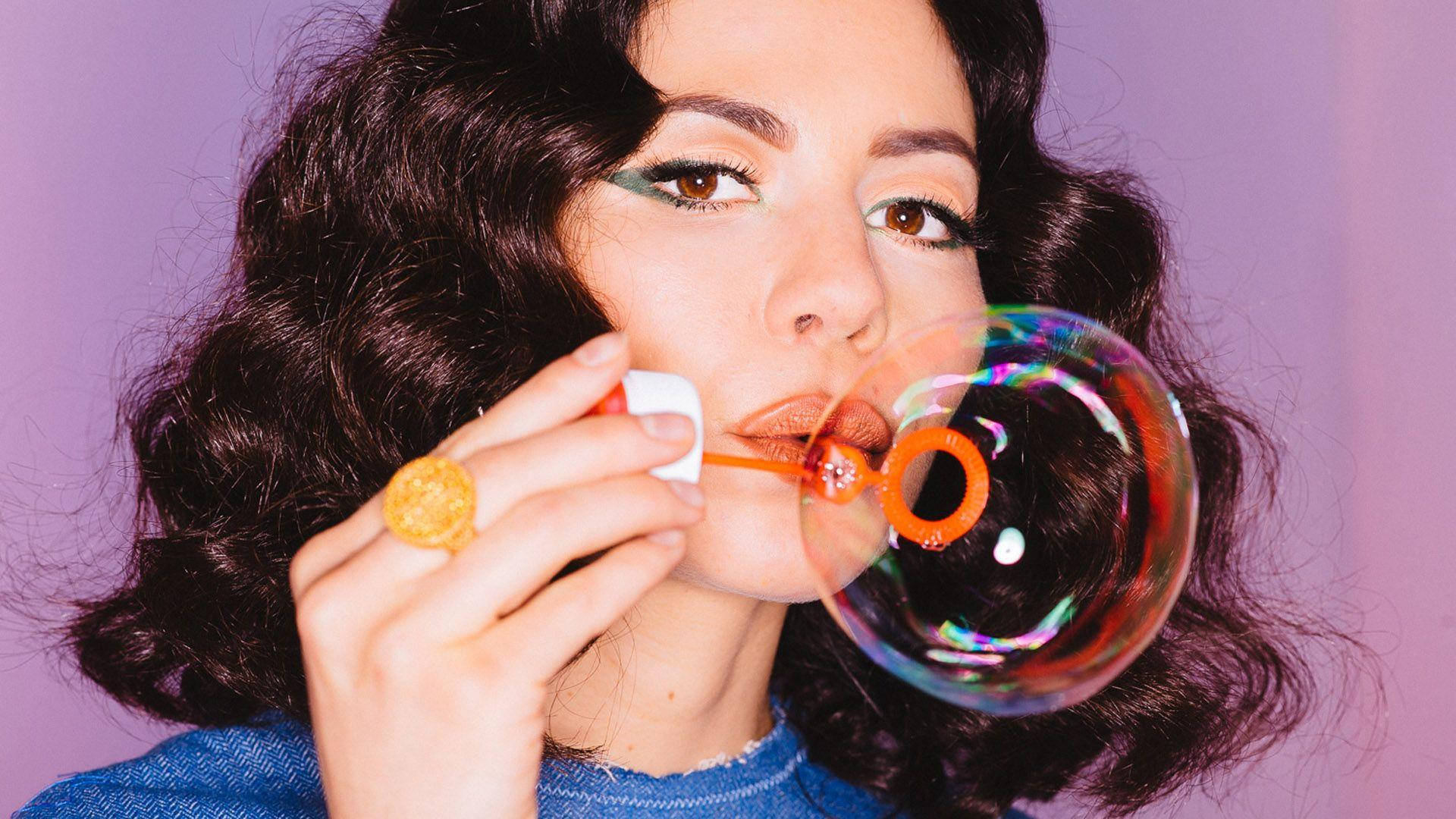 marina 桌布 壁紙 — 一位擁有深色捲髮的女性在柔和的紫色背景下吹出閃閃發光的彩虹泡泡。