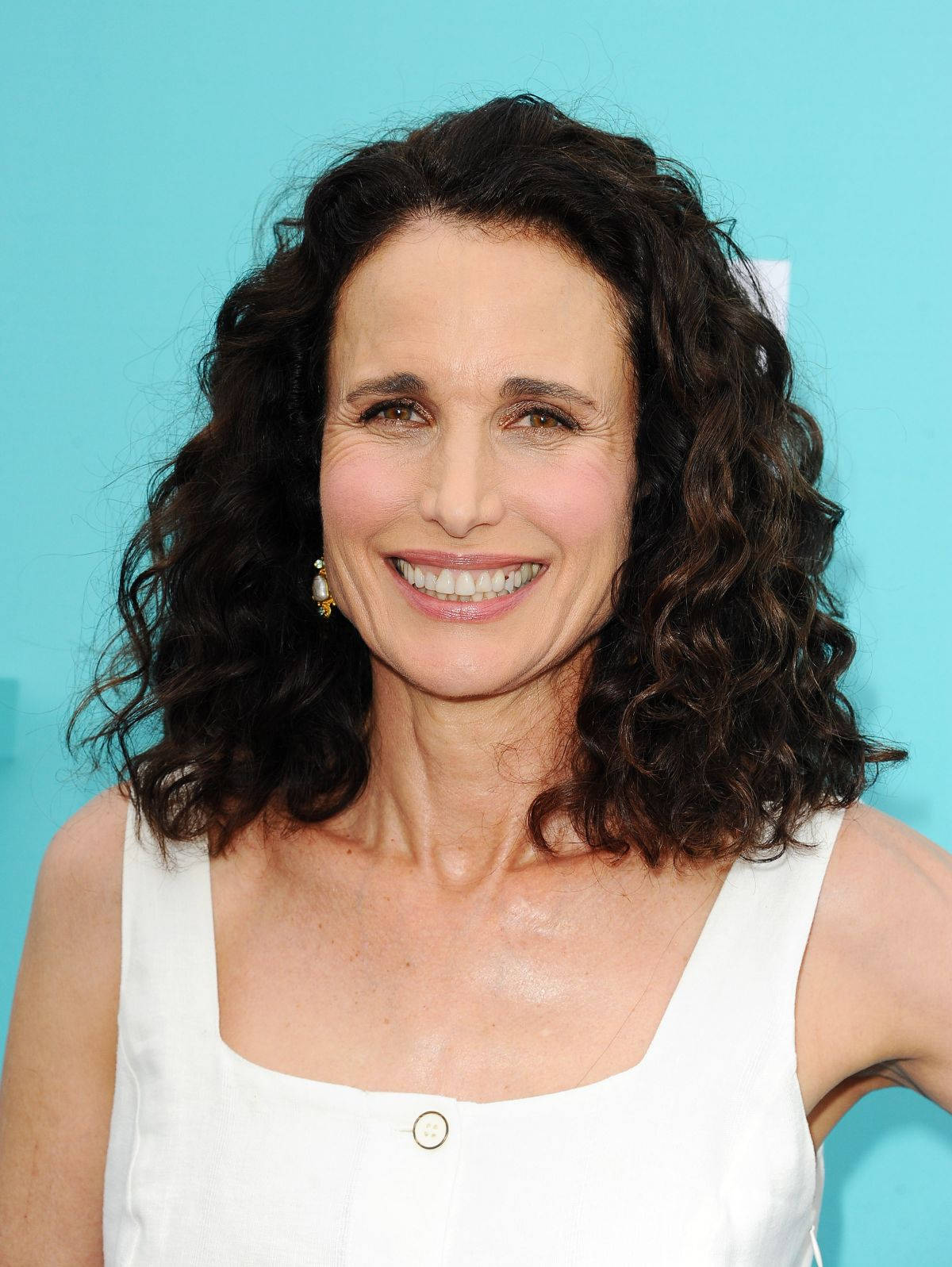 andie macdowell 桌布 壁紙 — 一位擁有深色捲髮的女性在純綠松色背景前穿著白色無袖上衣，溫暖地微笑。