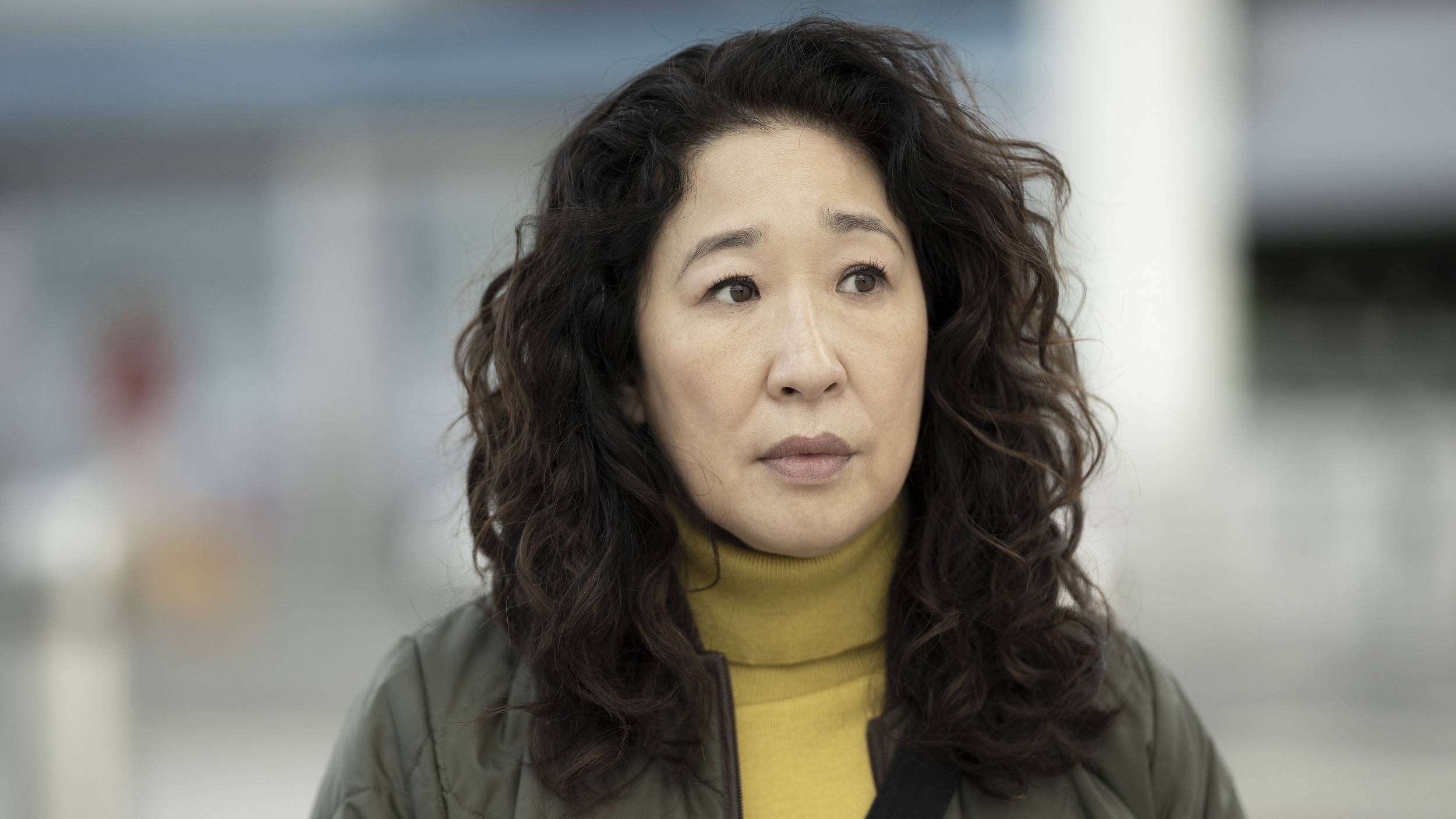killing eve 桌布 壁紙 — 一位擁有深色捲髮的女性穿著黃色高領毛衣，帶著沉思的表情看向鏡頭外。