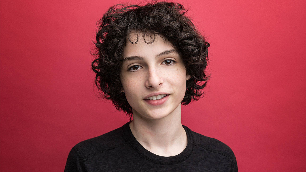 finn wolfhard 桌布 壁紙 — 一位擁有深色捲髮的年輕人對著純紅色攝影棚背景溫柔微笑。