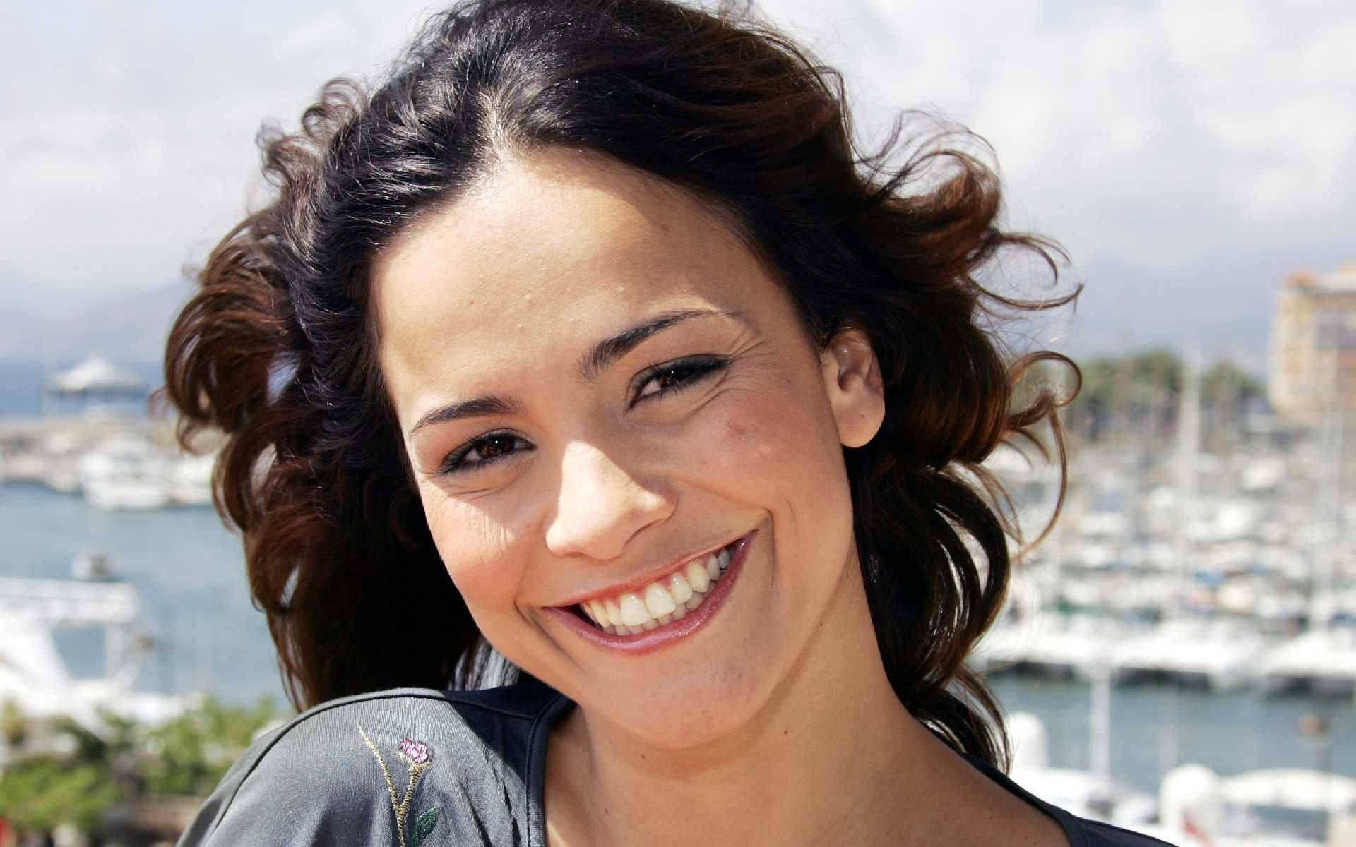 Celebrity 桌布 壁紙 — 一位擁有深色捲髮的燦爛女性，在充滿船隻和遠處建築物的陽光港口背景下燦爛微笑。
