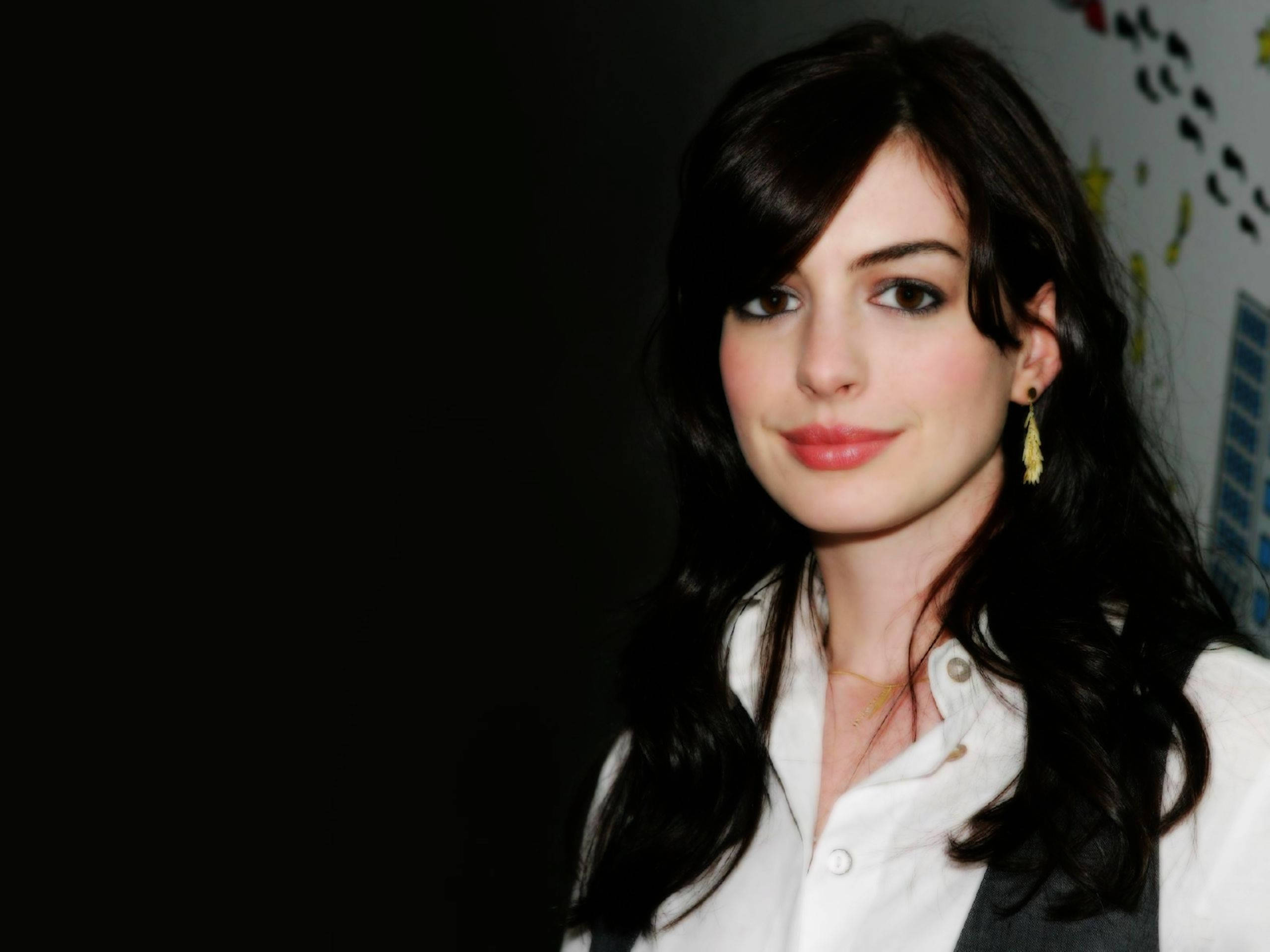 Anne Hathaway 桌布 壁紙 — 一位擁有深色波浪長髮、身穿白色襯衫的女性，在深色背景前微笑，身後有色彩繽紛的牆壁藝術。