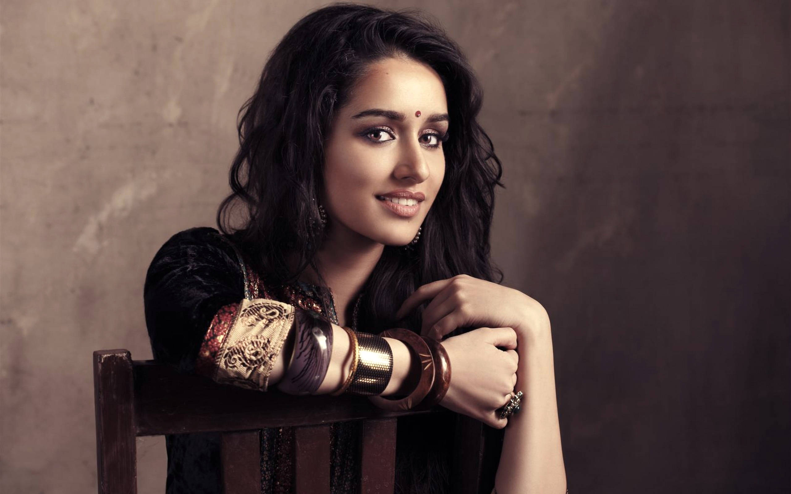 Shraddha Kapoor 桌布 壁紙 — 一位擁有深色波浪長髮和傳統珠寶的女性微笑著，將手臂擱在木椅上，靠著紋理豐富的棕色牆壁。