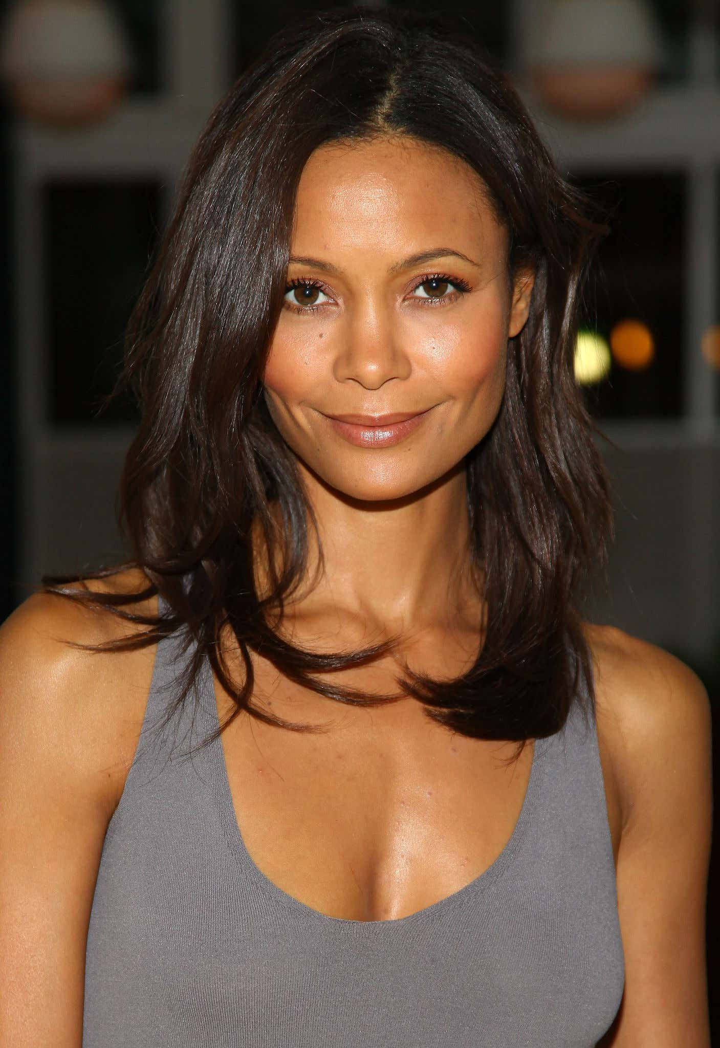 Actress HD 桌布 壁紙 — 一位擁有深色波浪長髮和溫柔微笑的女性，穿著灰色背心，背景柔和模糊。