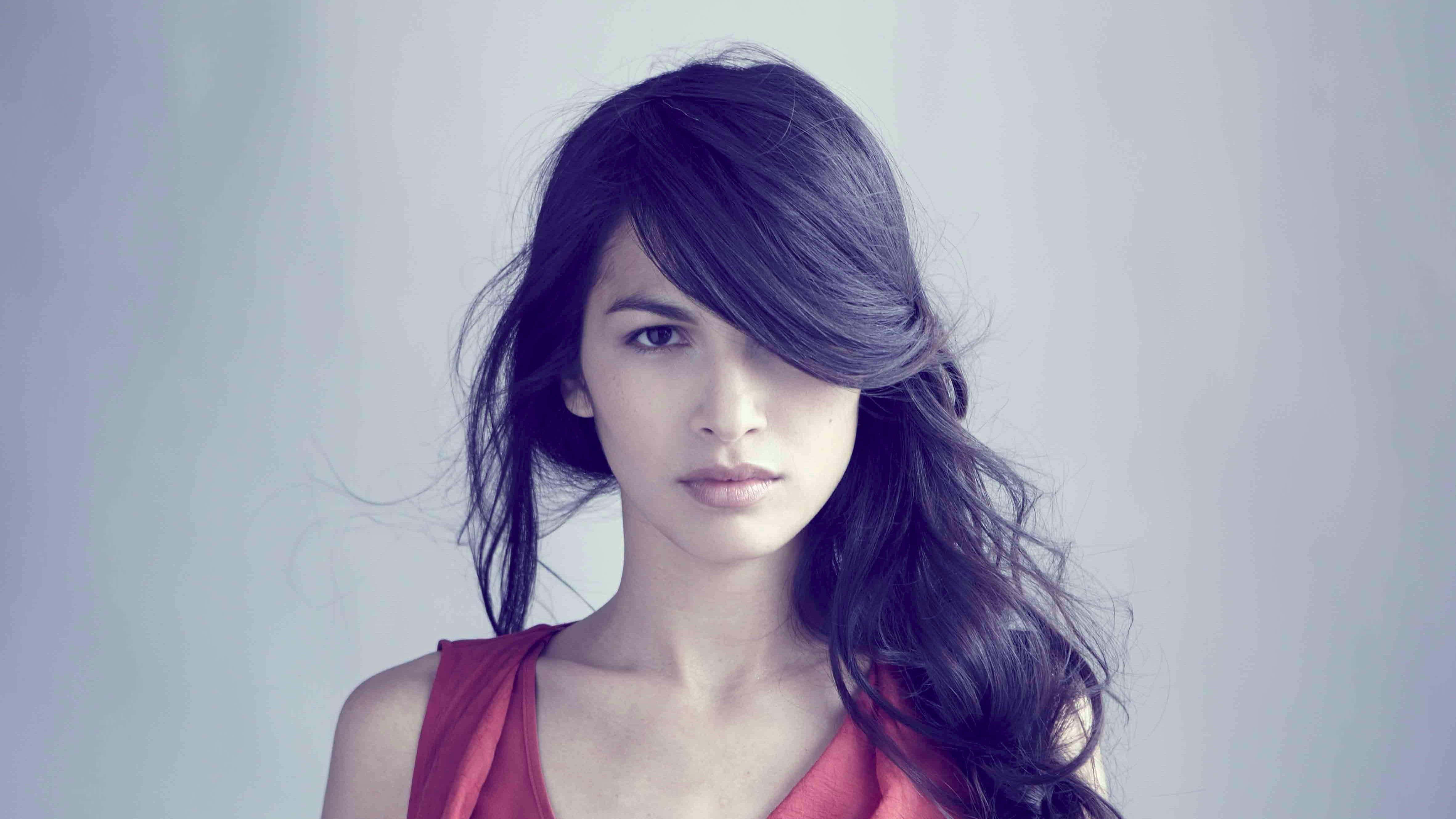 Elodie Yung 桌布 壁紙 — 一位擁有深色波浪長髮和紅色上衣的女性，在柔和的灰色背景下凝視著。