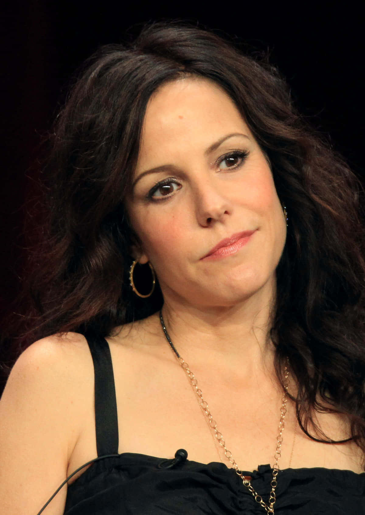 mary louise parker 桌布 壁紙 — 一位擁有深色波浪長髮和金色圓環耳環的女性，在深色背景下若有所思地凝視著。