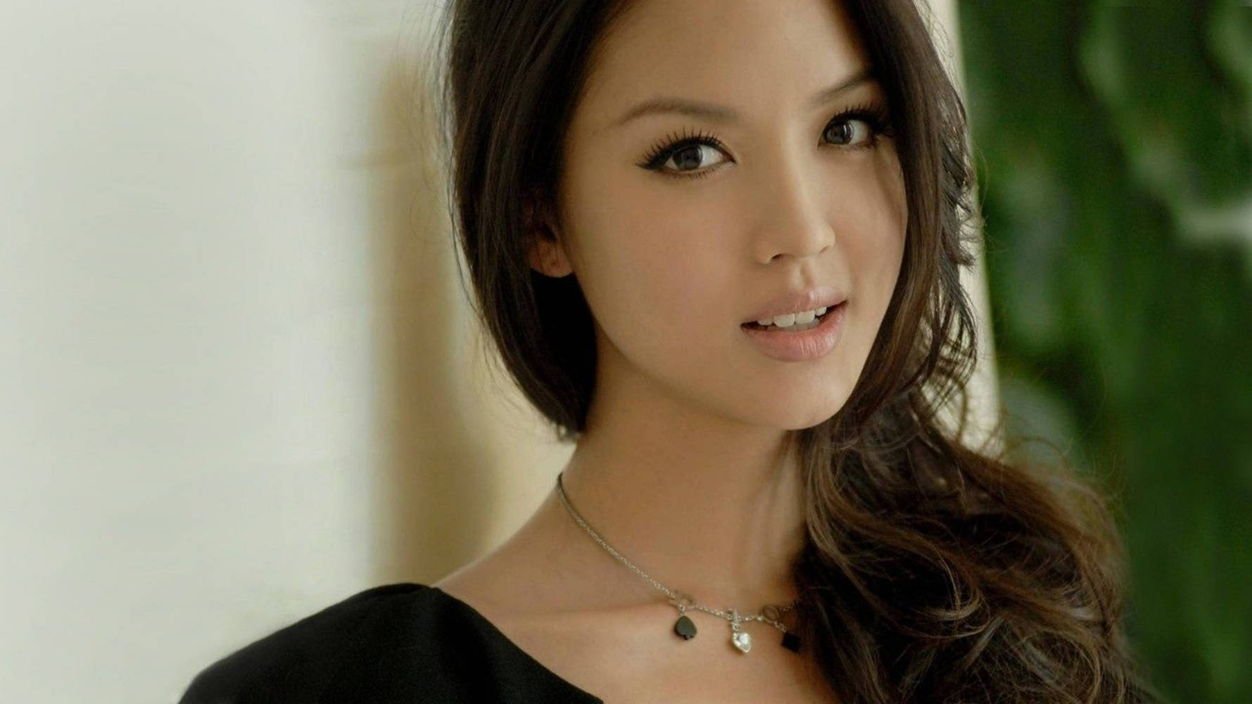 美女 桌布 壁紙 — 一位擁有深色波浪長髮和銀色項鍊的女性，在白色牆壁和綠色樹葉的襯托下溫柔地凝視著。