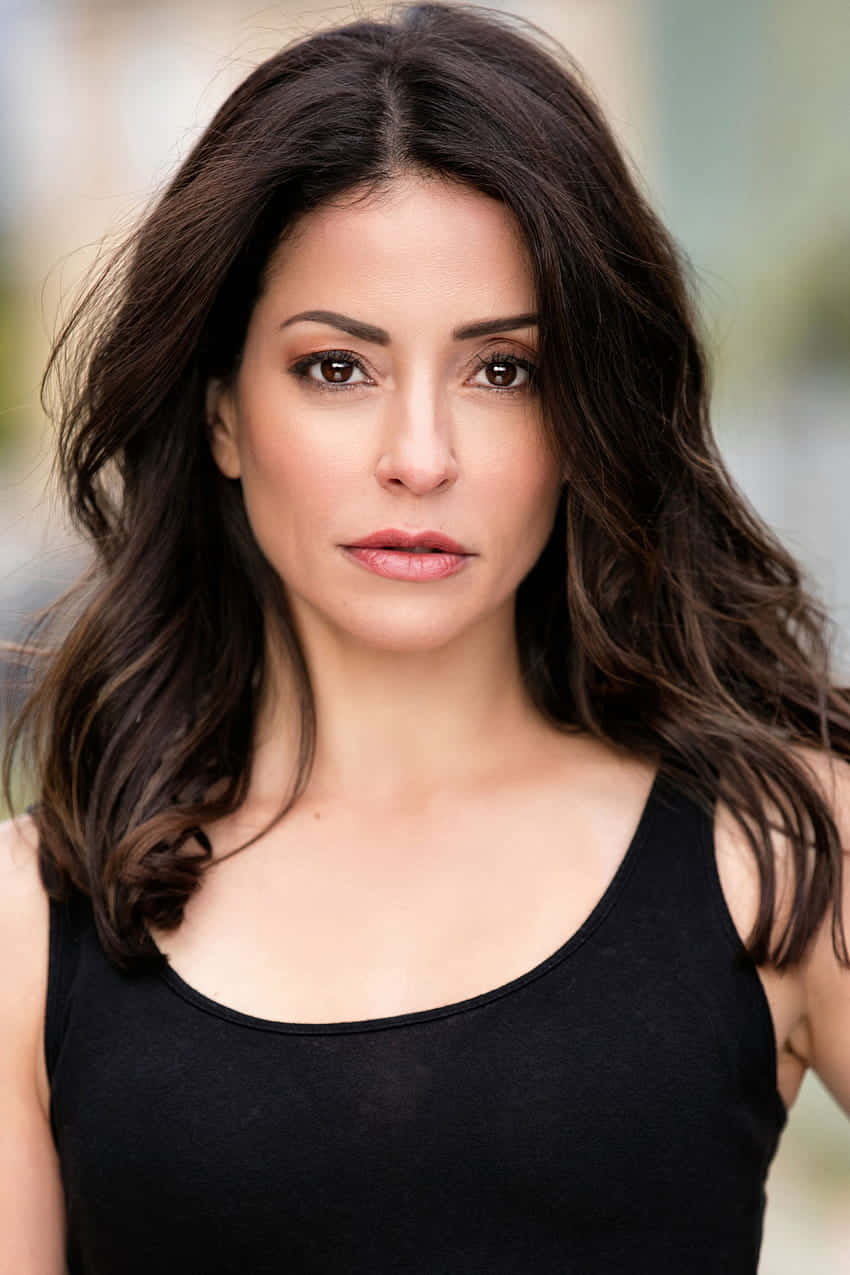Emmanuelle Vaugier 桌布 壁紙 — 一位擁有深色波浪長髮和銳利眼神的女性，在柔和模糊的背景前穿著黑色背心。