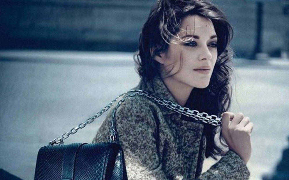 Actress HD 桌布 壁紙 — 一位擁有深色波浪長髮的女性，在冷色調的城市環境中，手握鏈條肩帶手袋，若有所思地凝視著。