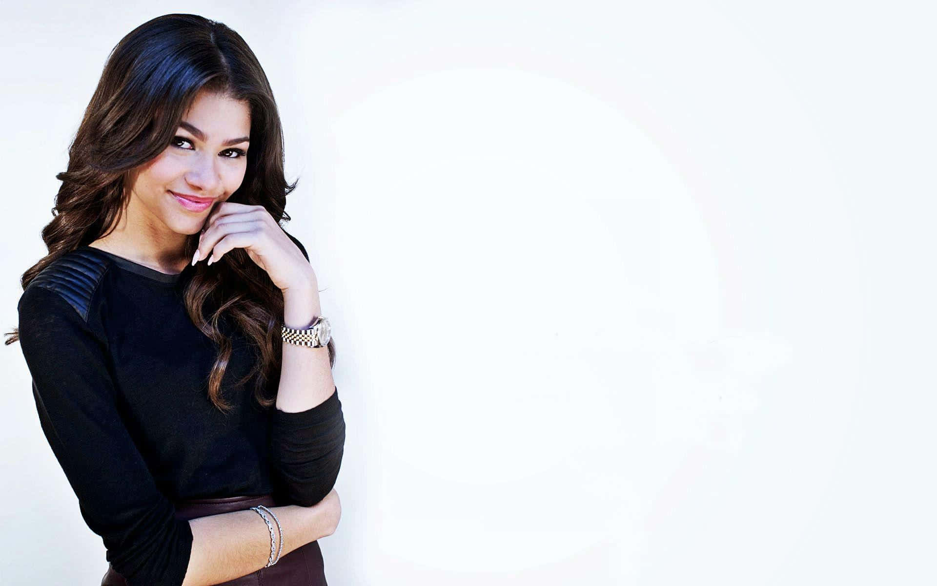 Zendaya 桌布 壁紙 — 一位擁有深色波浪長髮的女性，在明亮的白色背景上，將下巴擱在手上，溫柔地微笑。