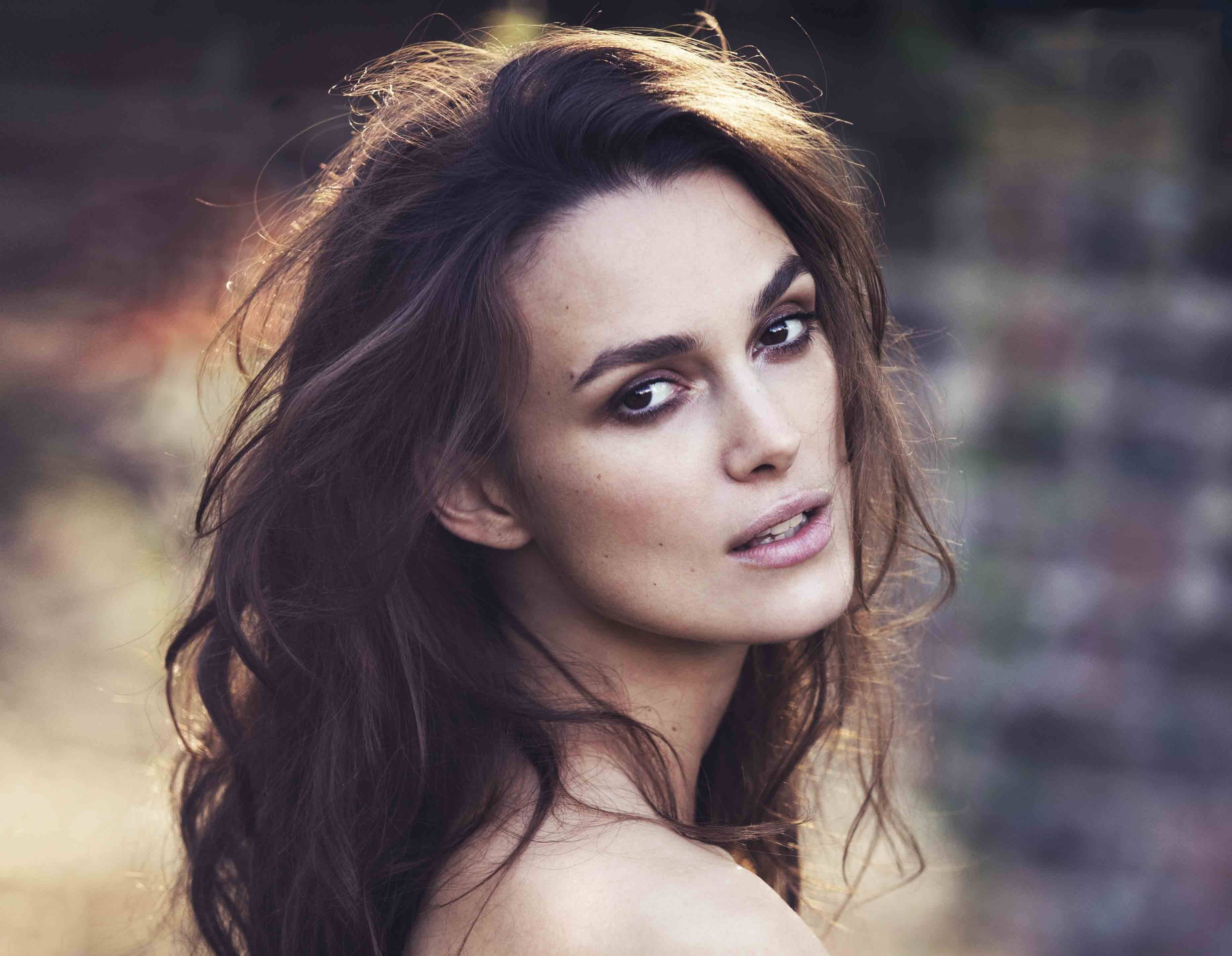 Keira Knightley 桌布 壁紙 — 一位擁有深色波浪長髮的女性，在柔和模糊的磚牆背景下專注地凝視鏡頭。