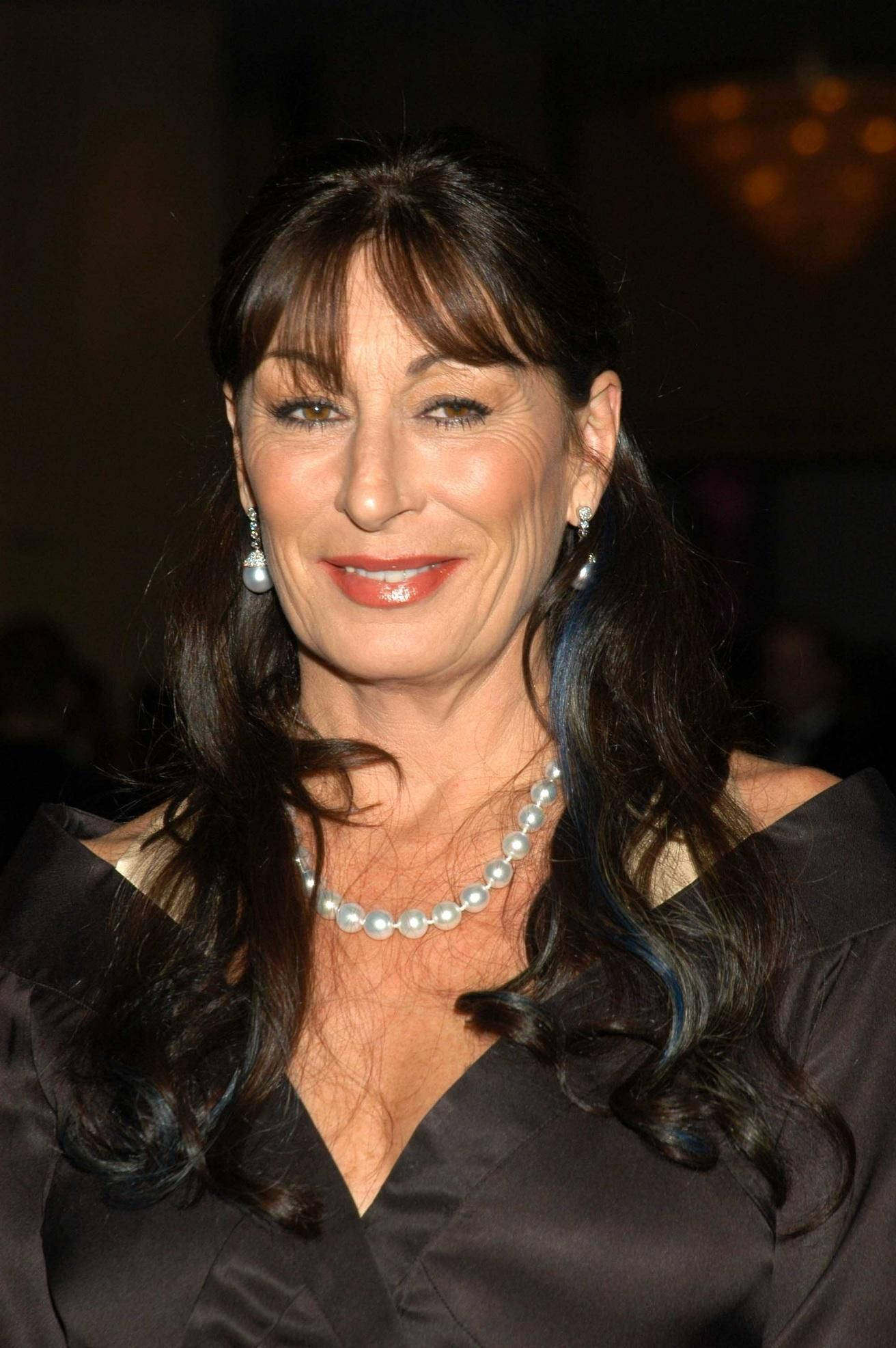 Celebrity 桌布 壁紙 — 一位擁有深色波浪長髮的女性，在柔和活動燈光下穿著黑色緞面連衣裙和珍珠首飾，溫暖地微笑。