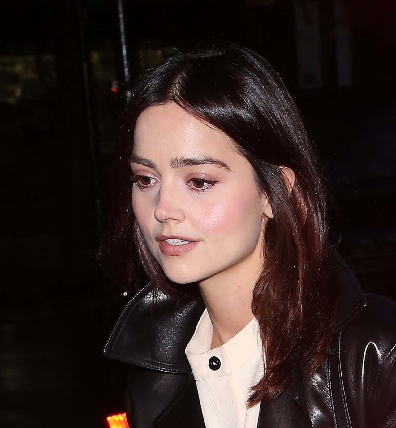 Jenna Coleman 桌布 壁紙 — 一位擁有深色波浪長髮的女性，在深色夜間背景下，身穿黑色皮外套內搭白色襯衫。