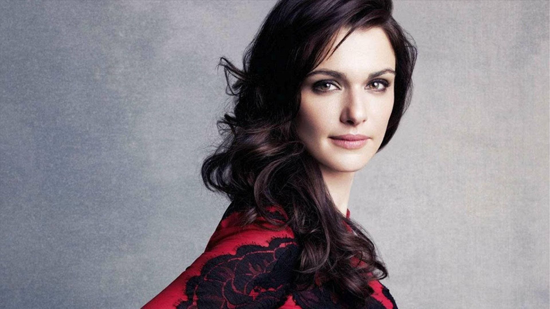 Rachel Weisz 桌布 壁紙 — 一位擁有深色波浪長髮的女性，穿著紅色花卉上衣，背景為灰色紋理背景。