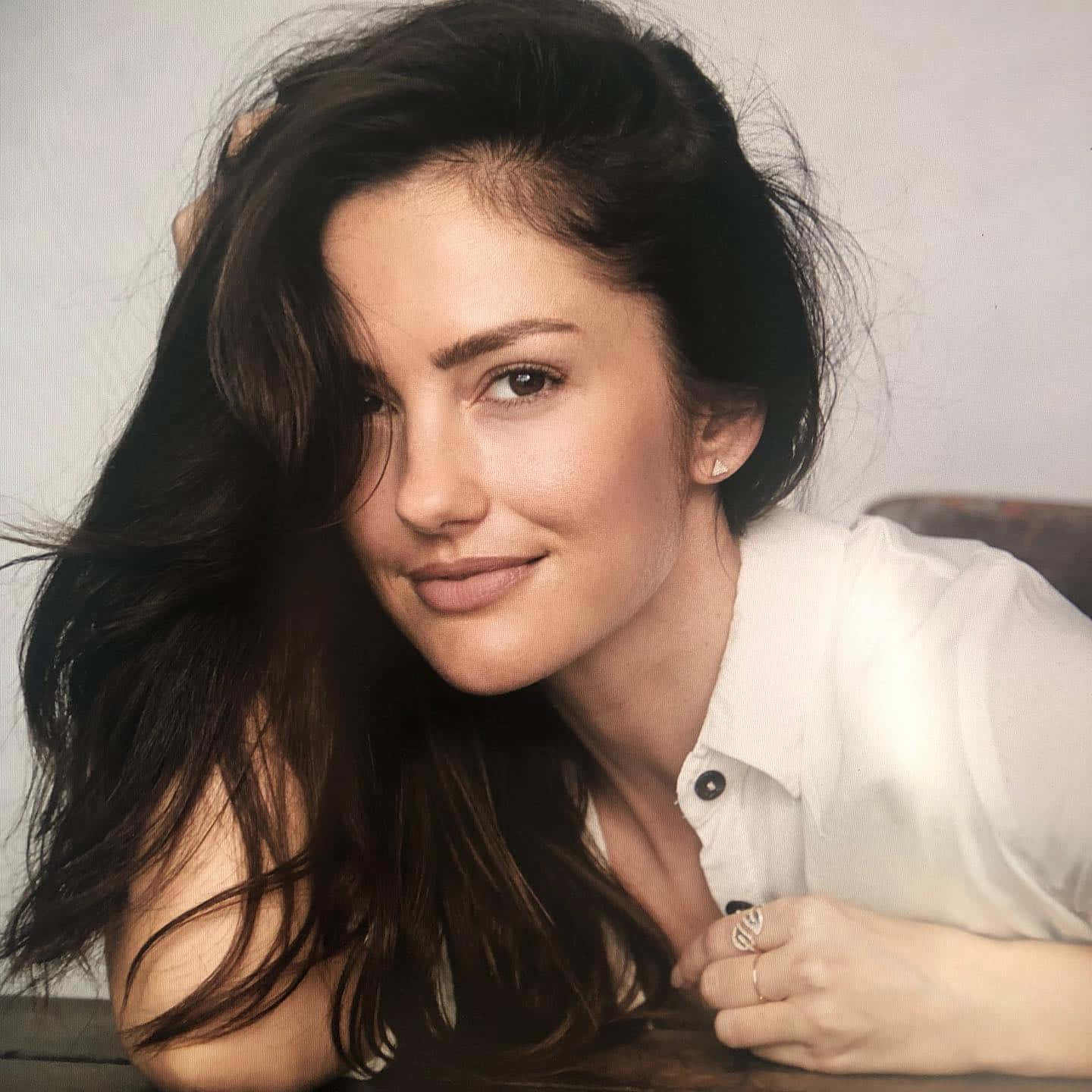 minka kelly 桌布 壁紙 — 一位擁有深色波浪長髮的女性，穿著白色襯衫，靠在木質表面上溫柔微笑。