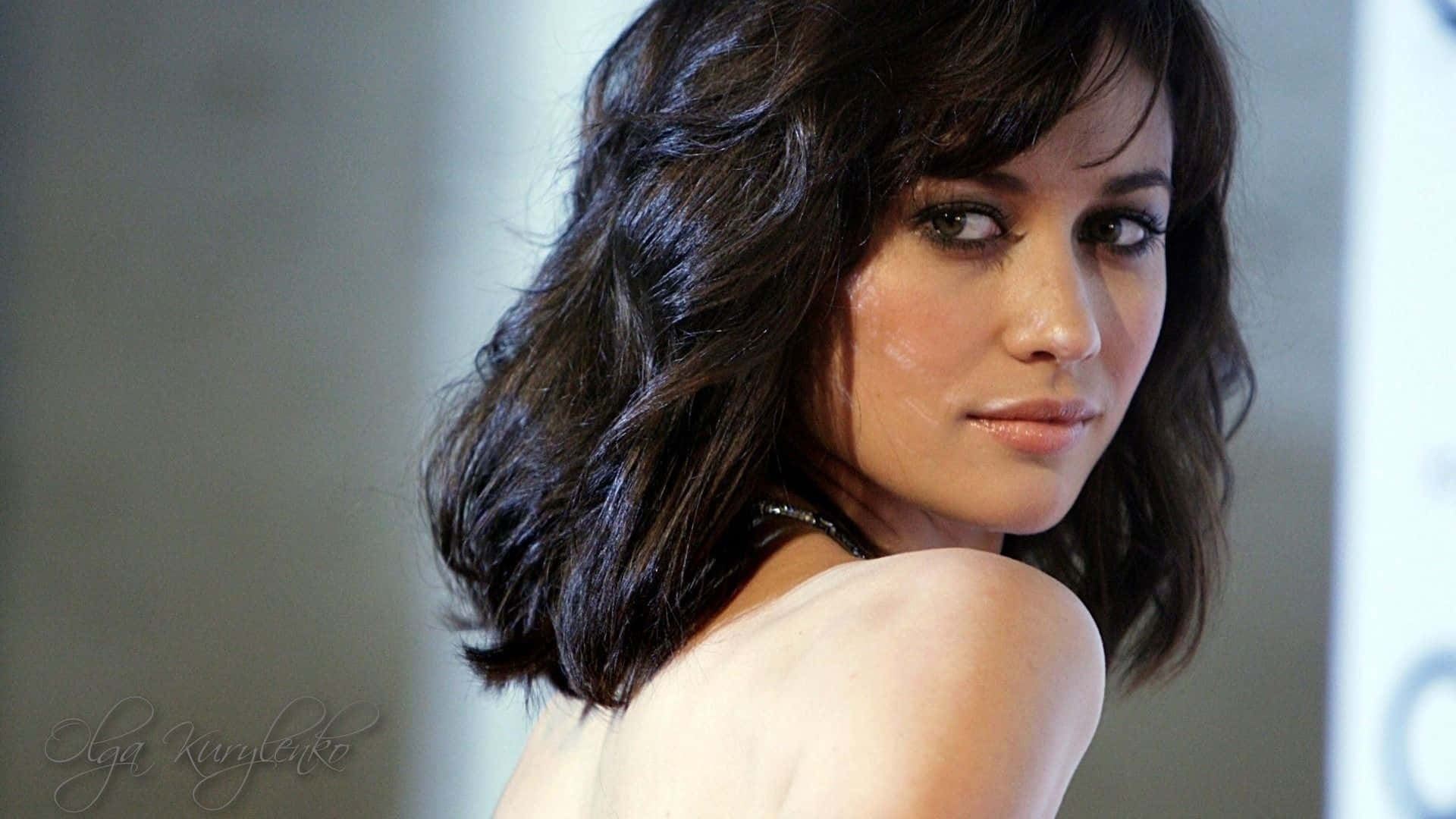 Olga Kurylenko 桌布 壁紙 — 一位擁有深色波浪長髮的女性，身穿白色上衣，溫柔地回頭望向肩膀後方。