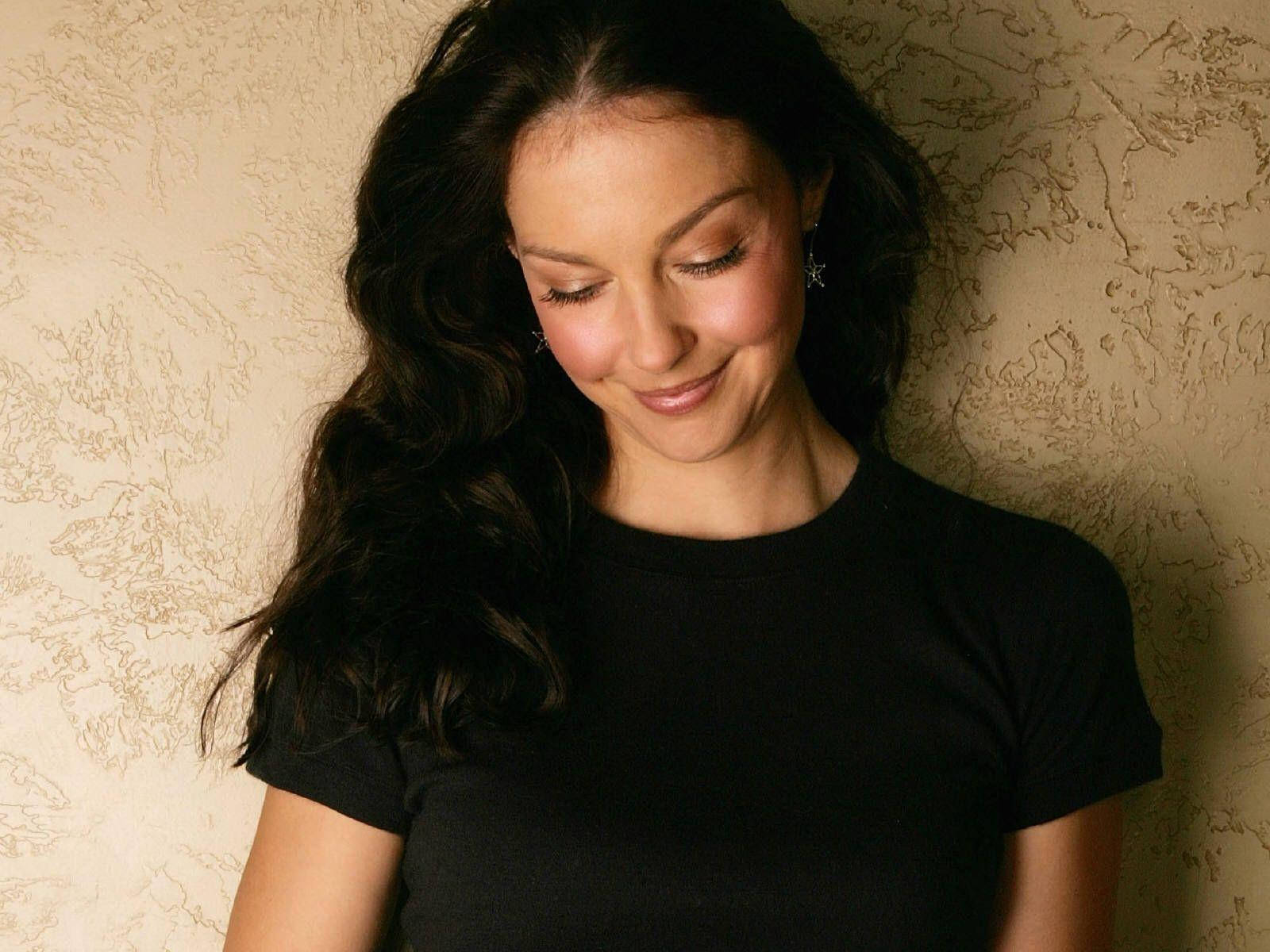 Ashley Judd 桌布 壁紙 — 一位擁有深色波浪長髮的女性，靠著質感米色牆壁，穿著黑色T恤，溫柔微笑。